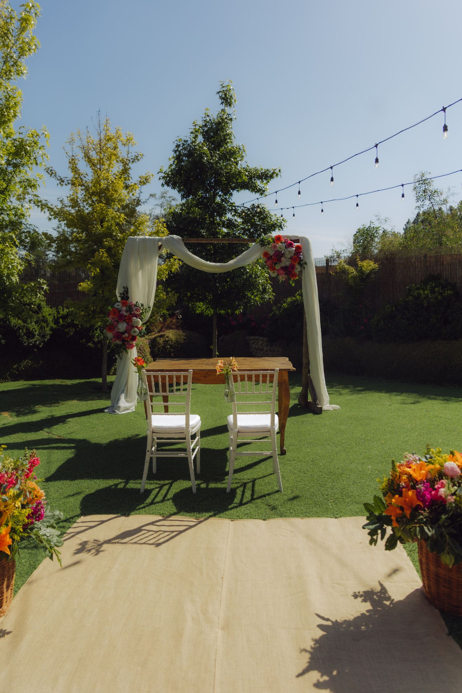 Outdoor wedding altar with white draping, floral accents, and string lights in a garden.