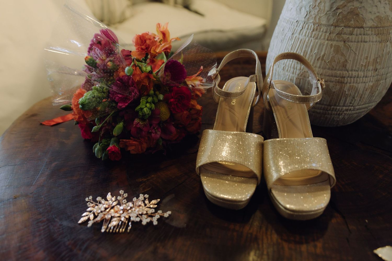 Gold glitter bridal heels, floral hair comb, and vibrant bouquet on a wooden table.