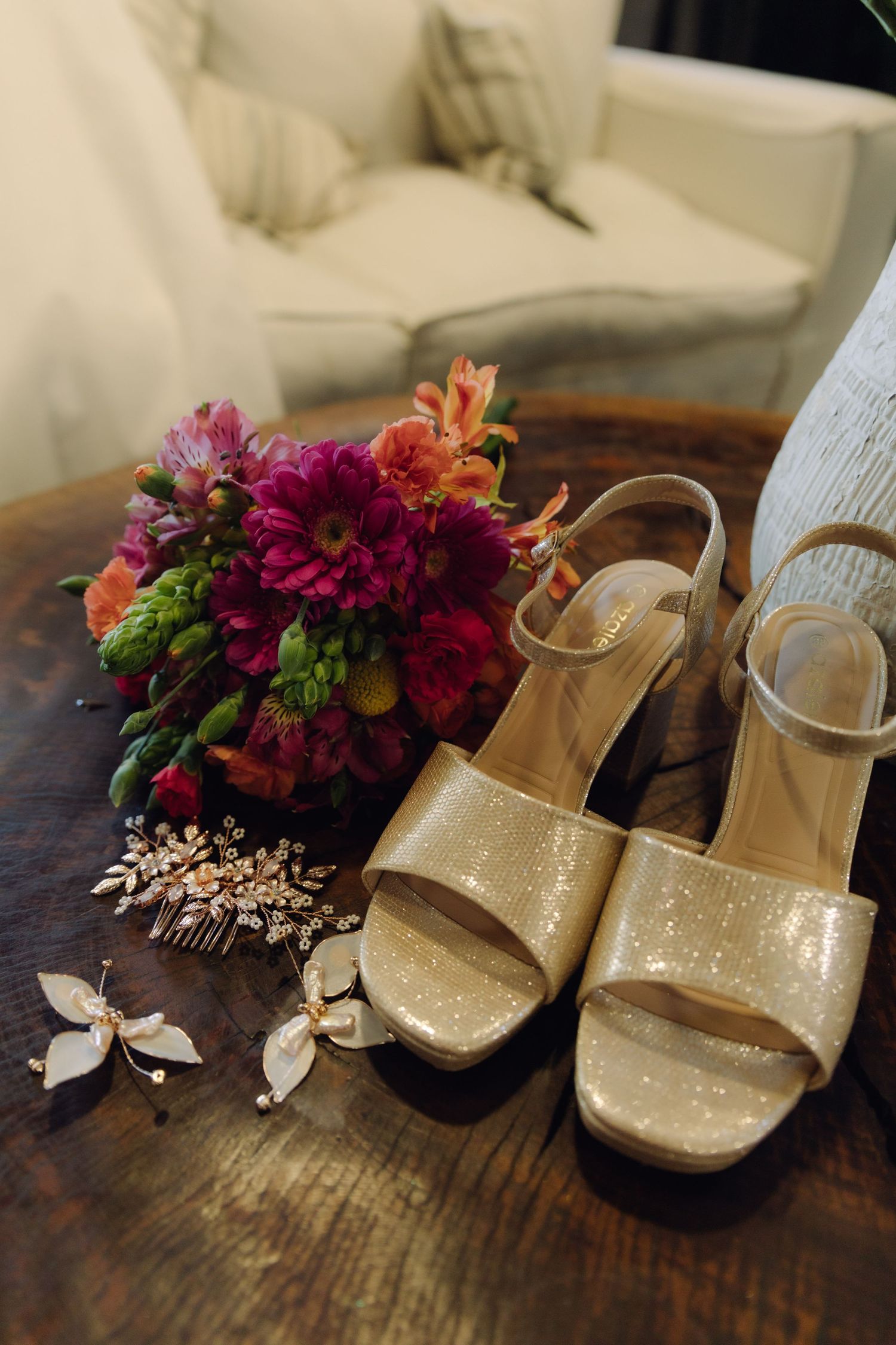 Gold glitter bridal heels, butterfly earrings, hair comb, and colorful bouquet on wood table.