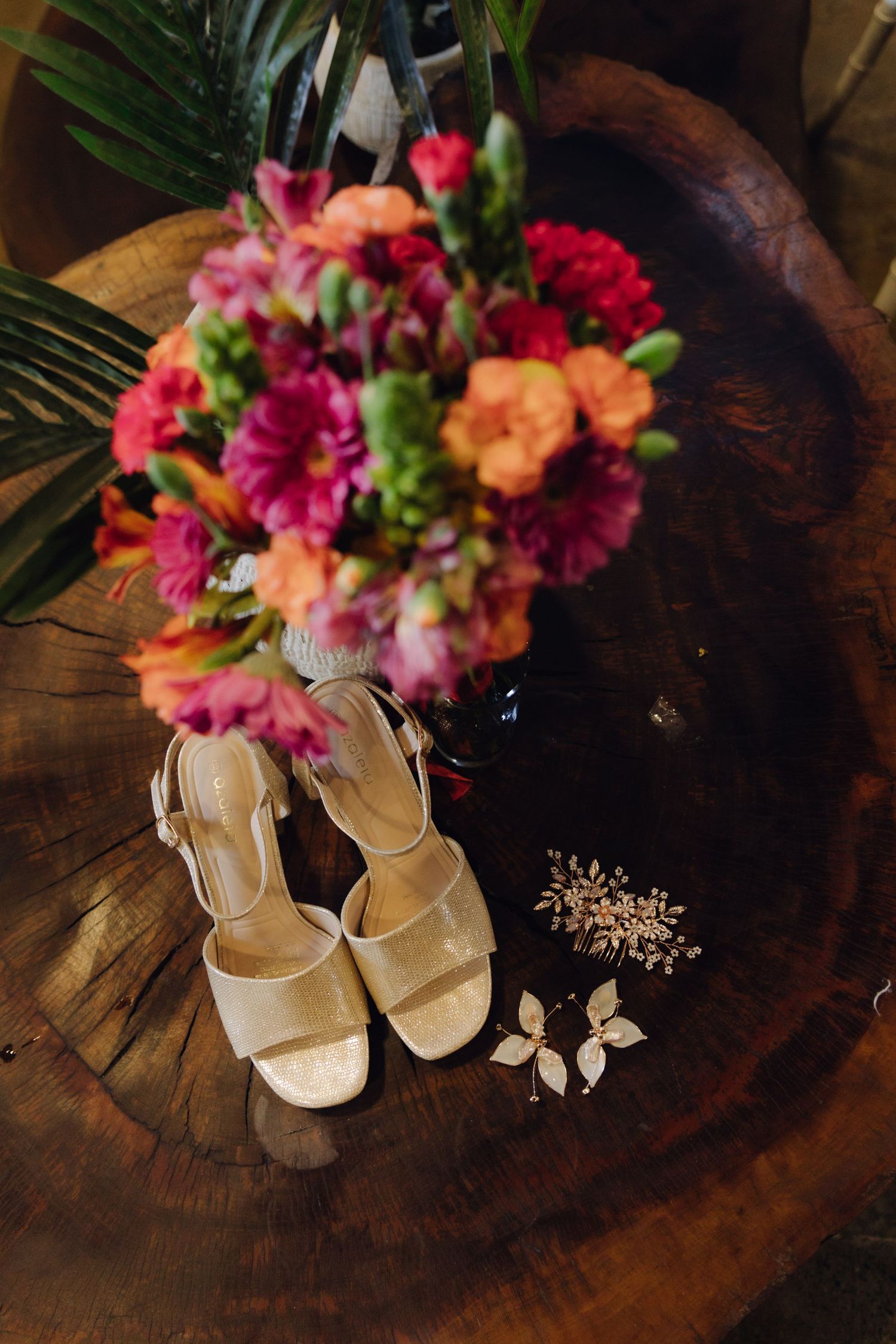 Gold bridal sandals, floral hair accessories, and vibrant pink and orange bouquet on wood.
