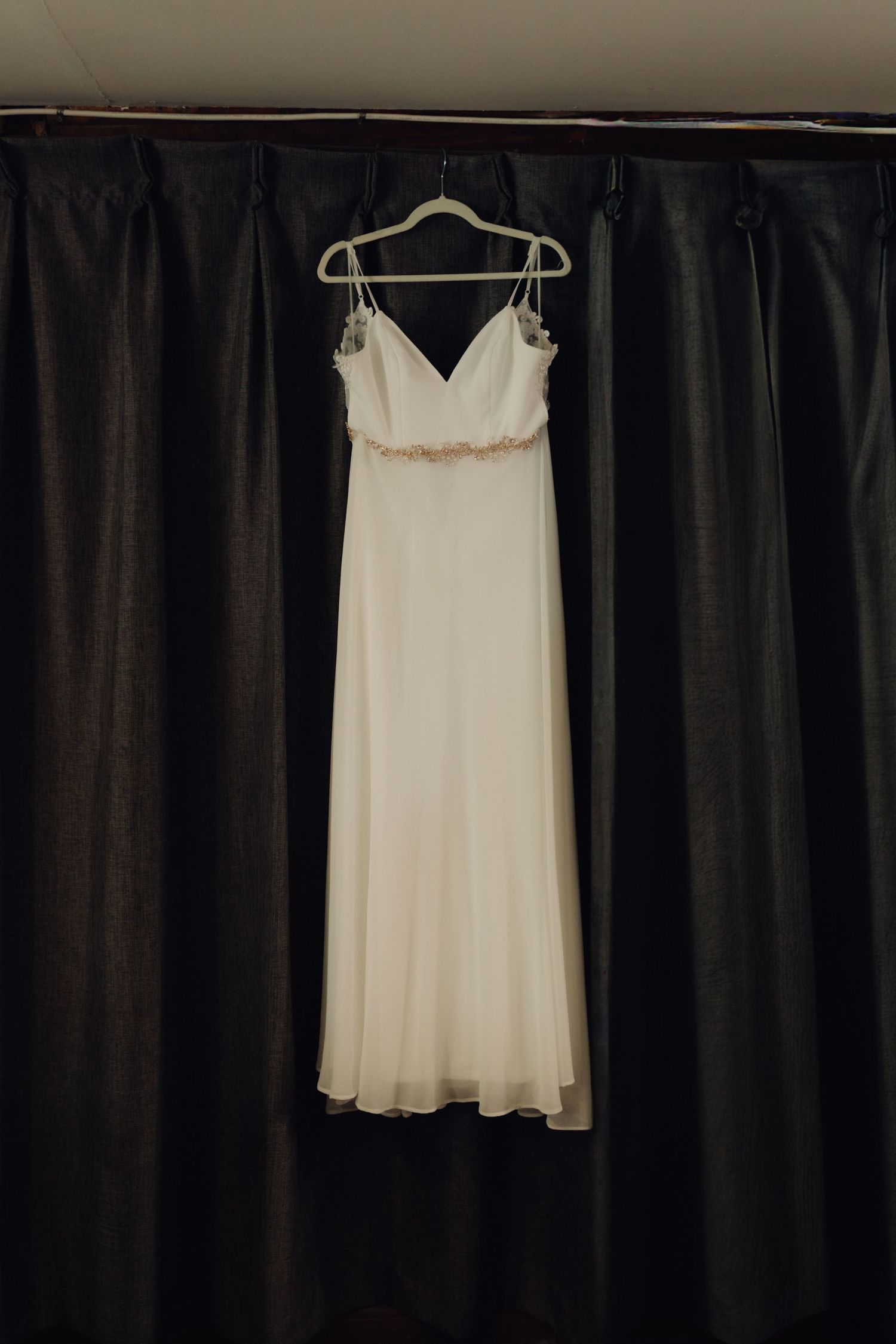 Elegant white bridal gown with spaghetti straps hanging on a hanger against dark curtains.