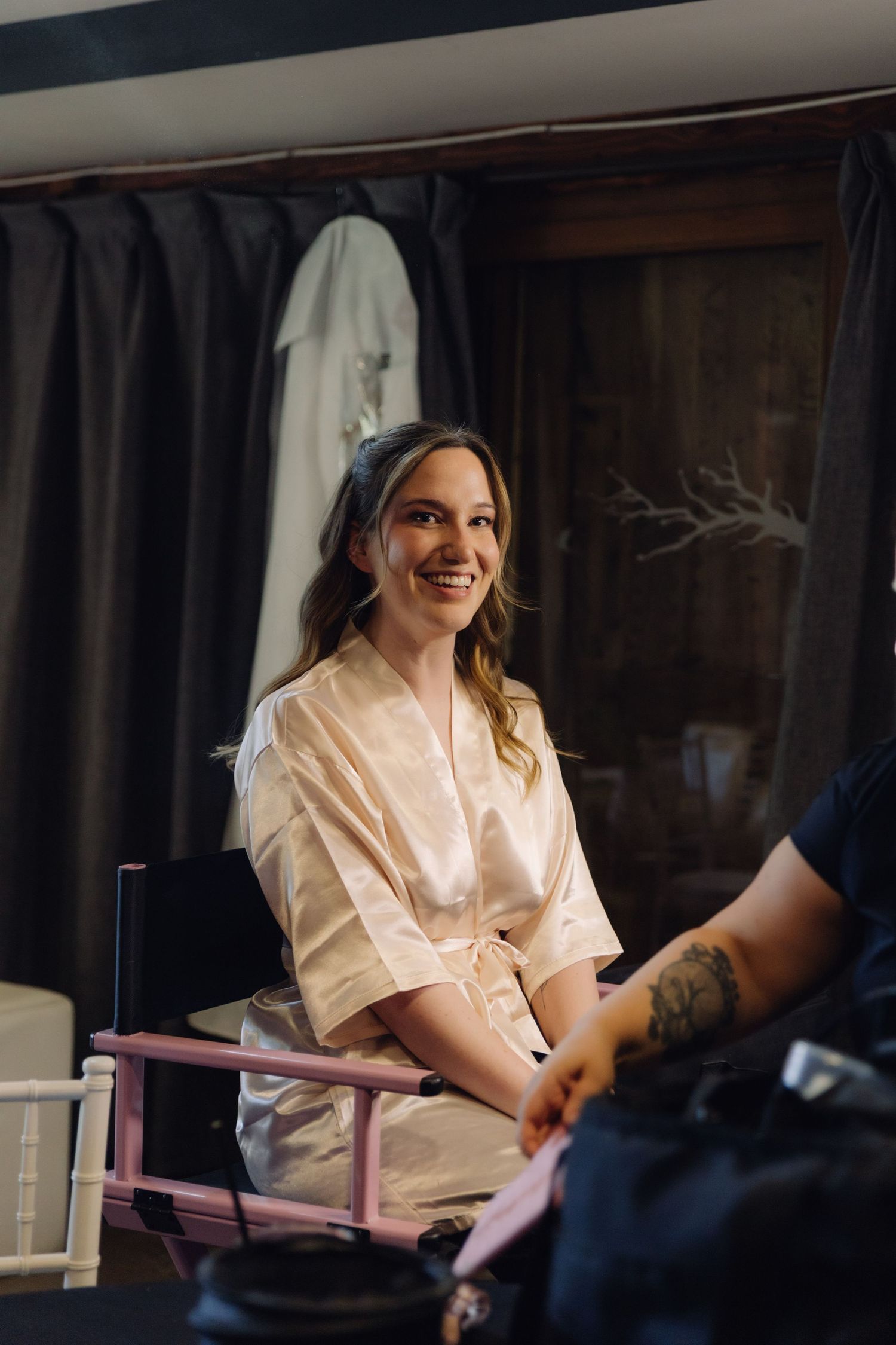 Smiling bride in a satin robe sitting in a pink makeup chair during bridal preparations.