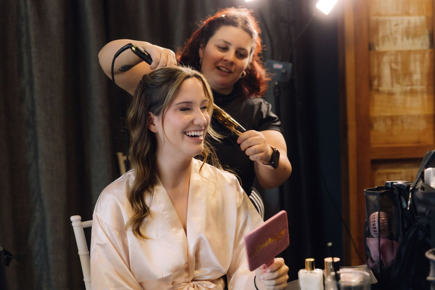 Bride laughing joyfully while hairstylist styles her hair during wedding day preparations.