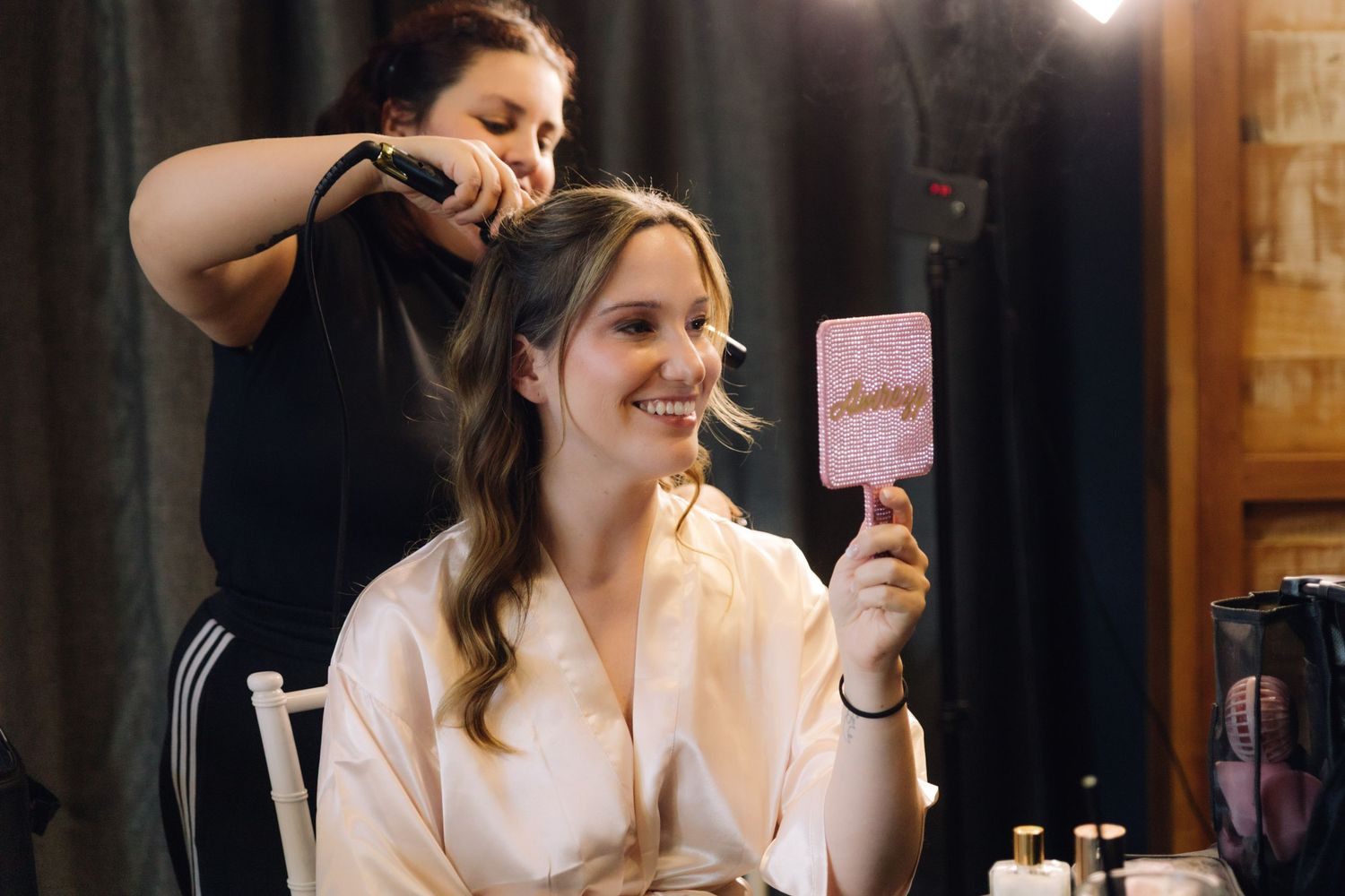 Bride smiling and holding a pink mirror as her hair is styled before the wedding ceremony.
