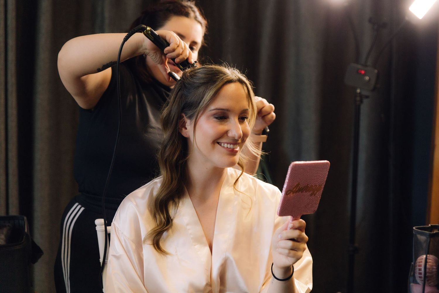 Bride smiling at her reflection in a pink mirror while getting her hair done for her wedding.