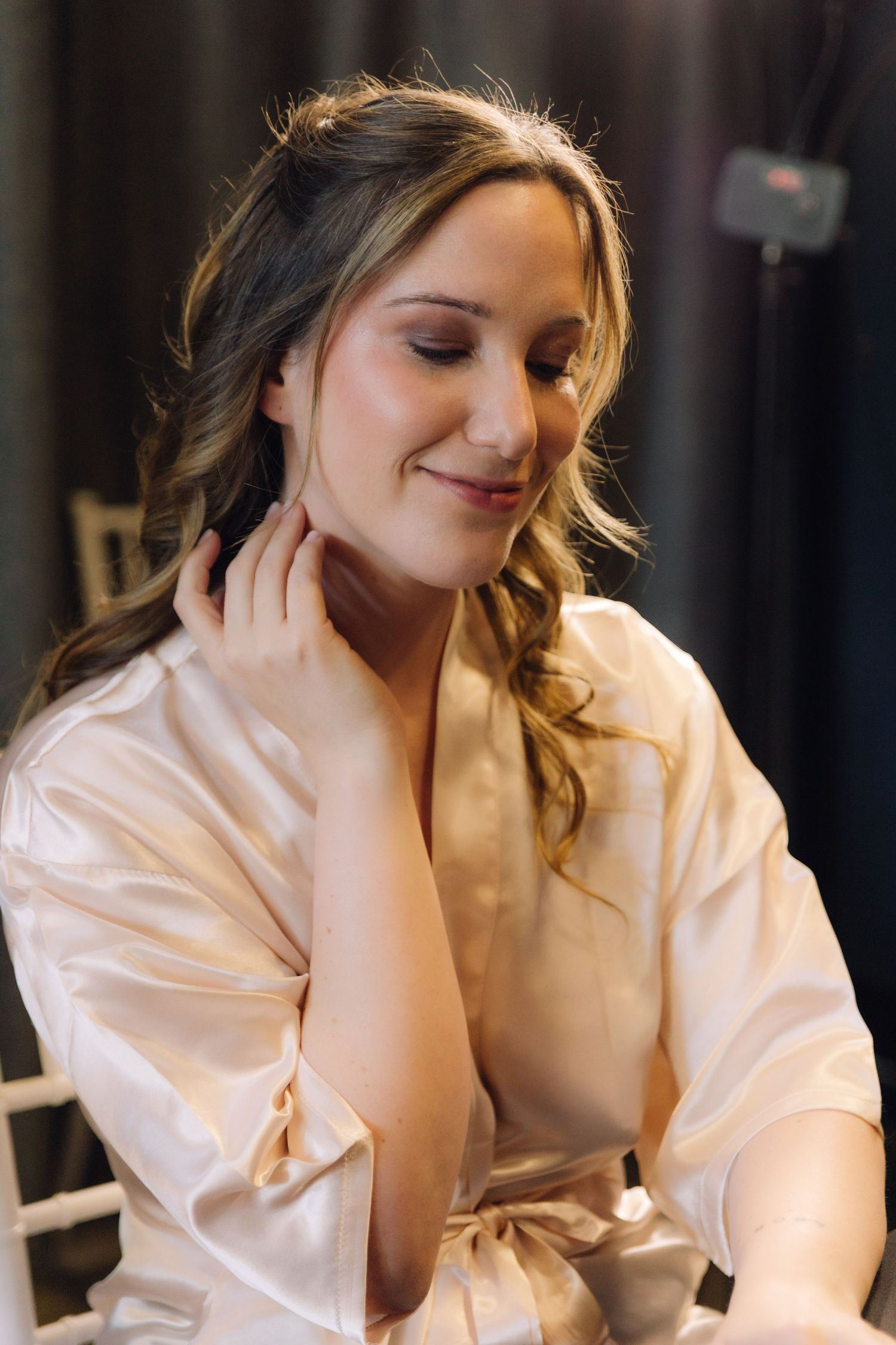Beautiful bride in a satin robe touching her neck and smiling softly after hair and makeup.
