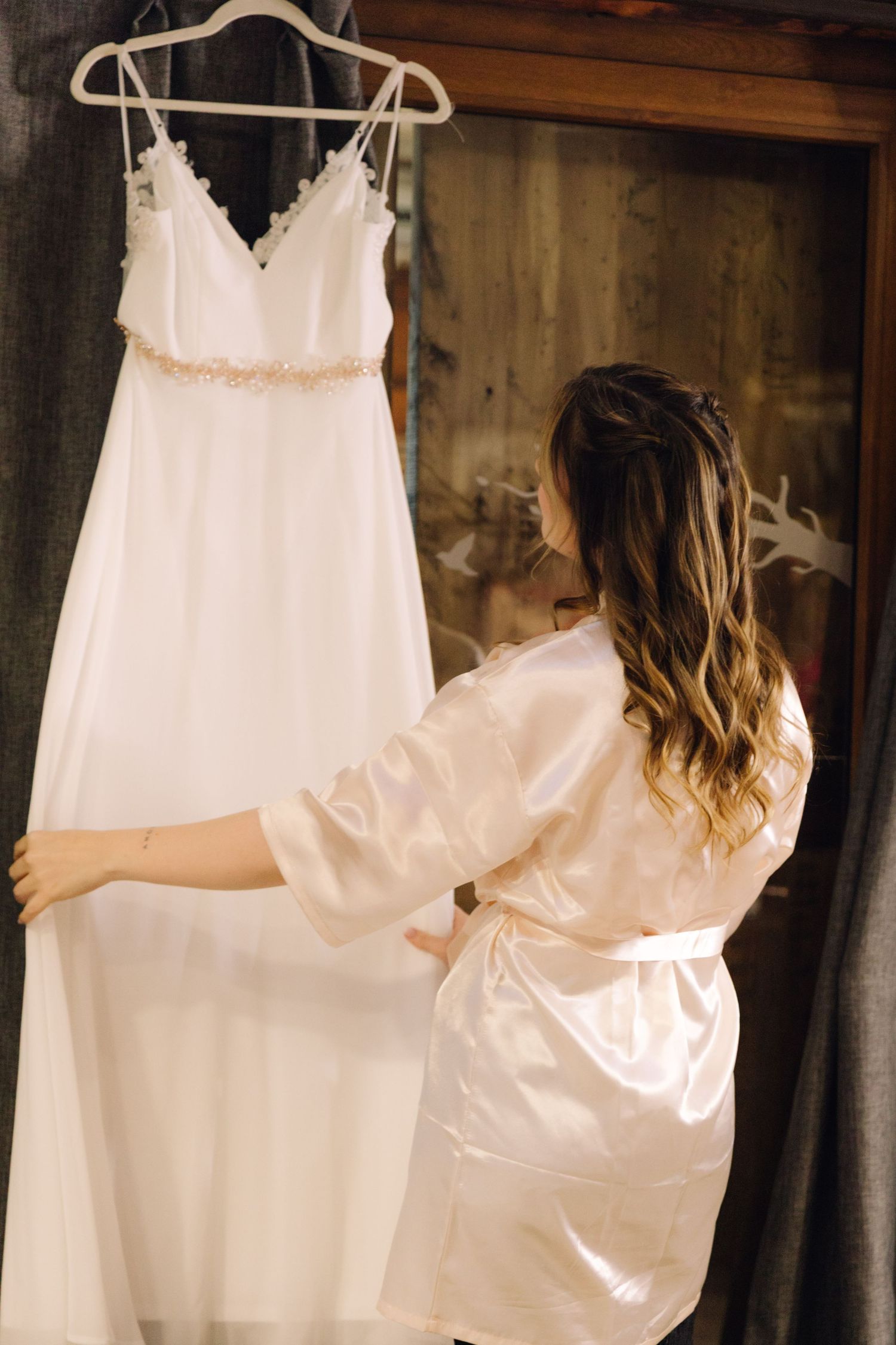Bride in satin robe touching her elegant white wedding gown hanging on a rustic wooden door.