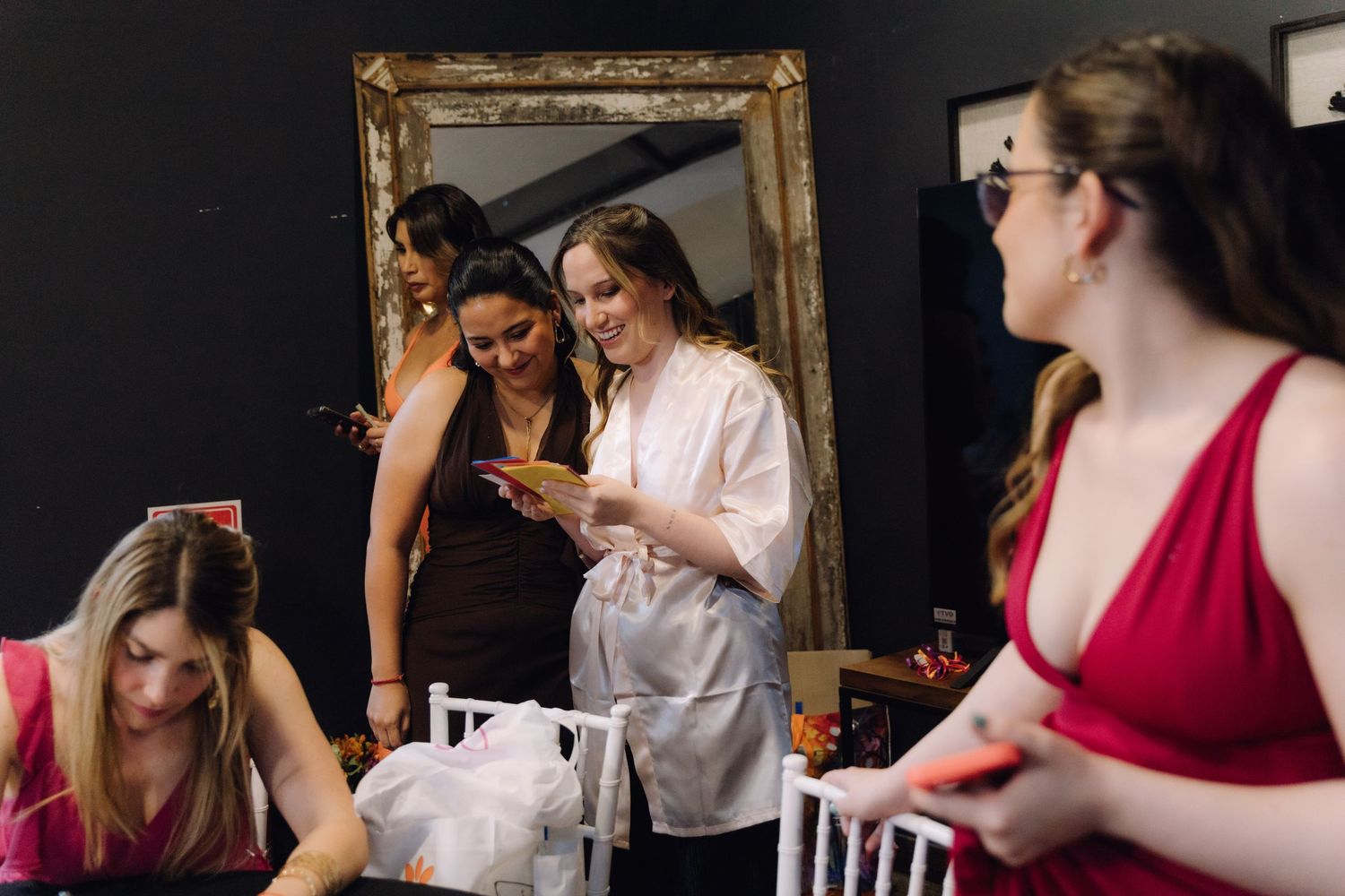 Bride in white satin robe smiling while reading a card surrounded by bridesmaids in red dresses.