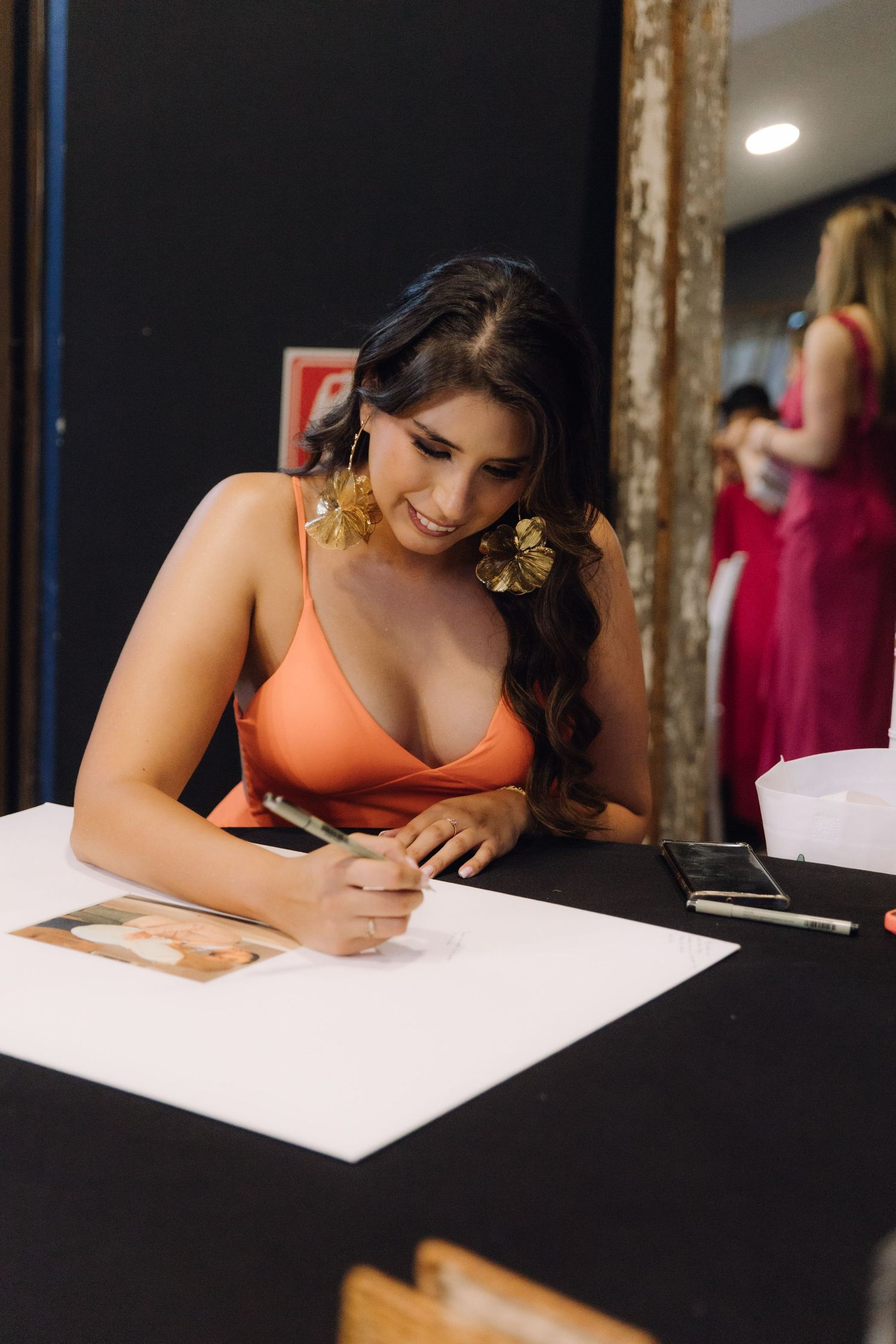 Elegant woman in orange dress with gold earrings signing a guestbook at a wedding reception table.