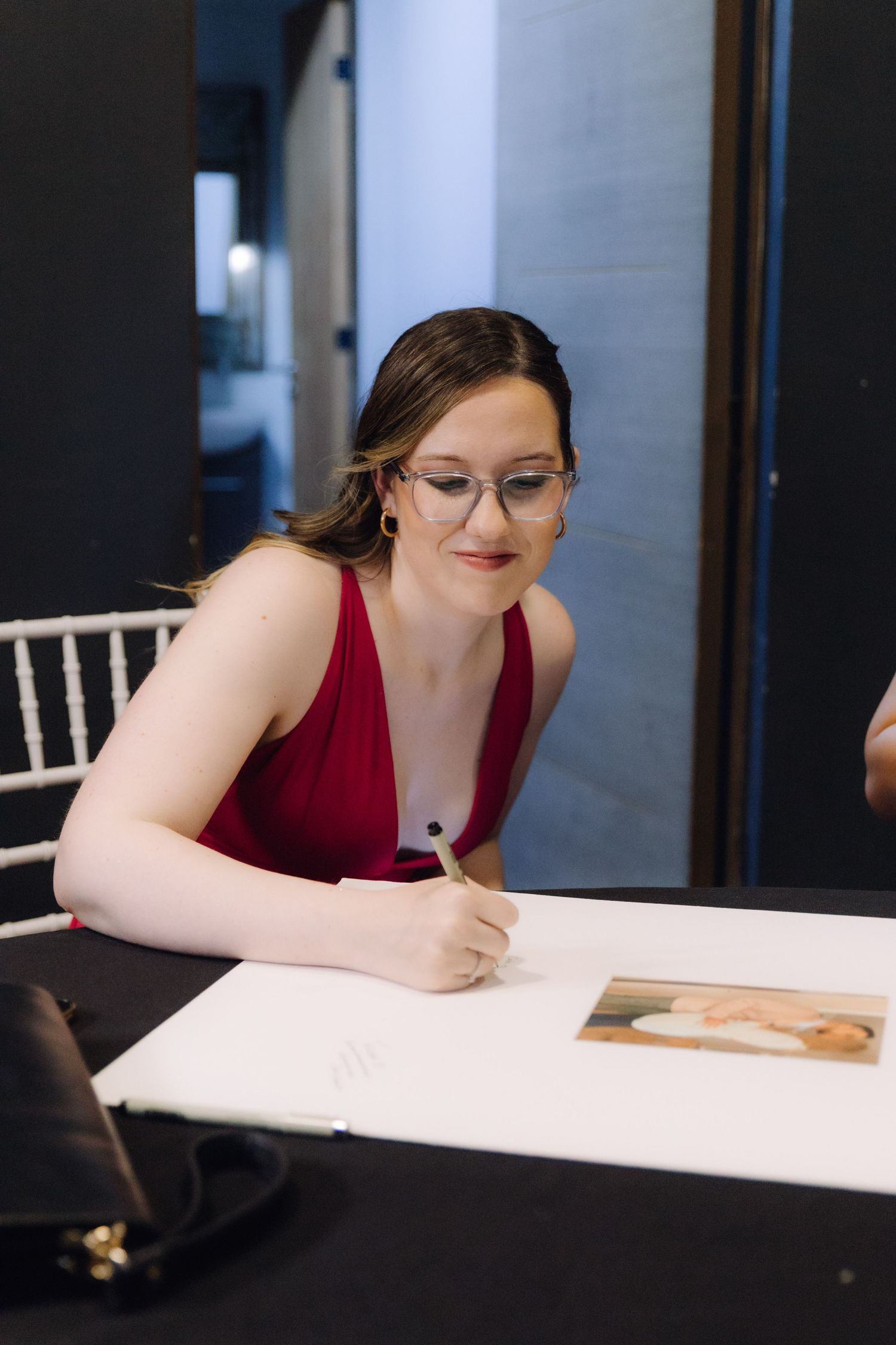 Young woman wearing glasses and red dress writing in a wedding guestbook at a reception table.