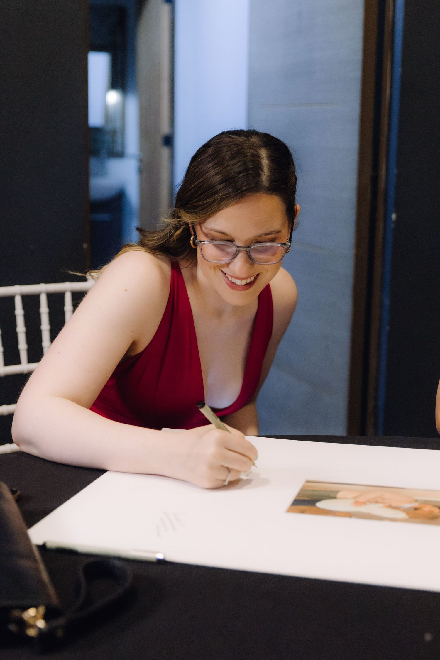 Smiling woman in red dress and glasses signing a wedding guestbook at a black table.