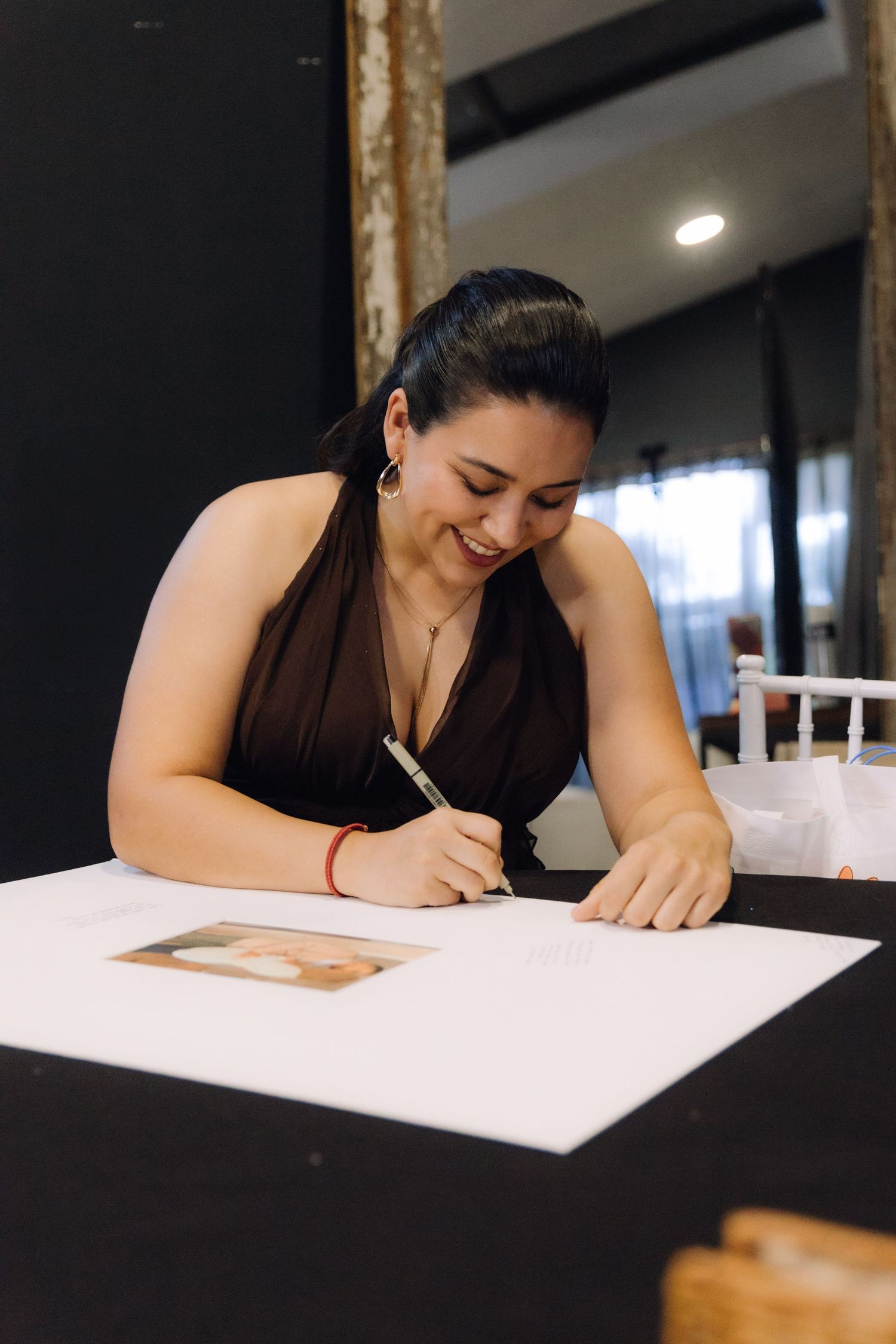 Smiling woman in black halter dress signing a wedding guestbook at a white table.