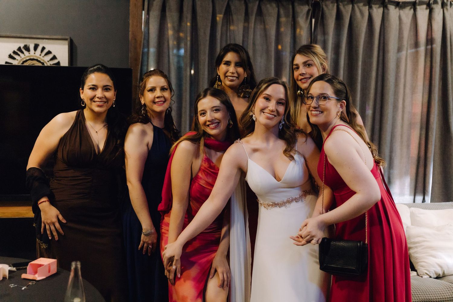 Bride in white dress poses joyfully with bridesmaids in red and black gowns.