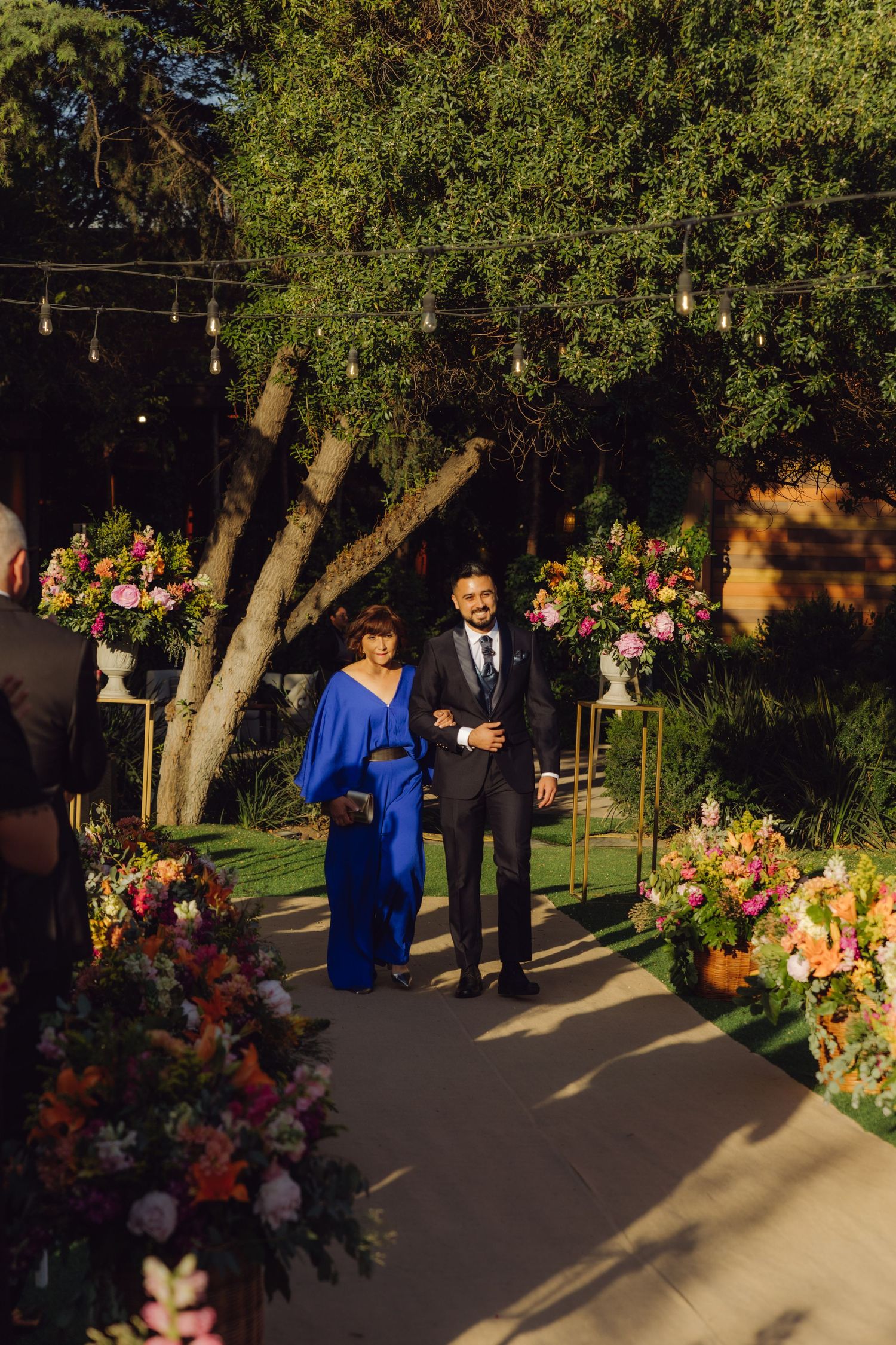 Groom and mother walking down a floral-lined wedding aisle surrounded by colorful arrangements.