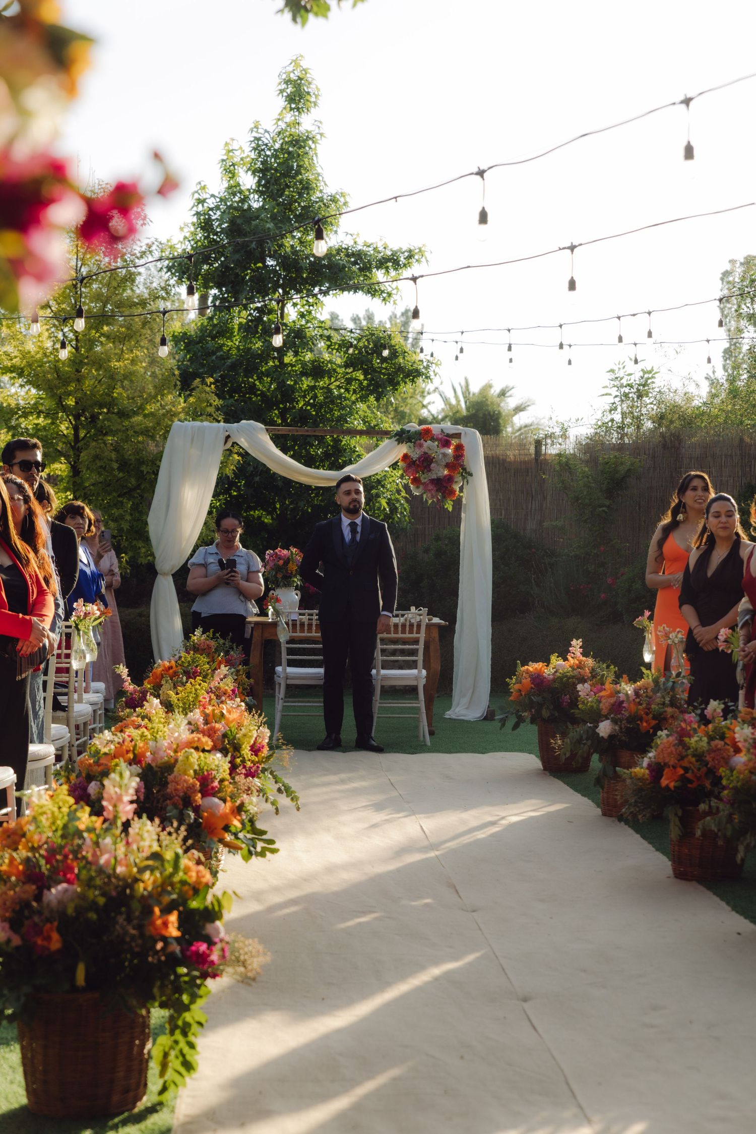 Groom standing at a floral wedding altar with white draping and string lights overhead.