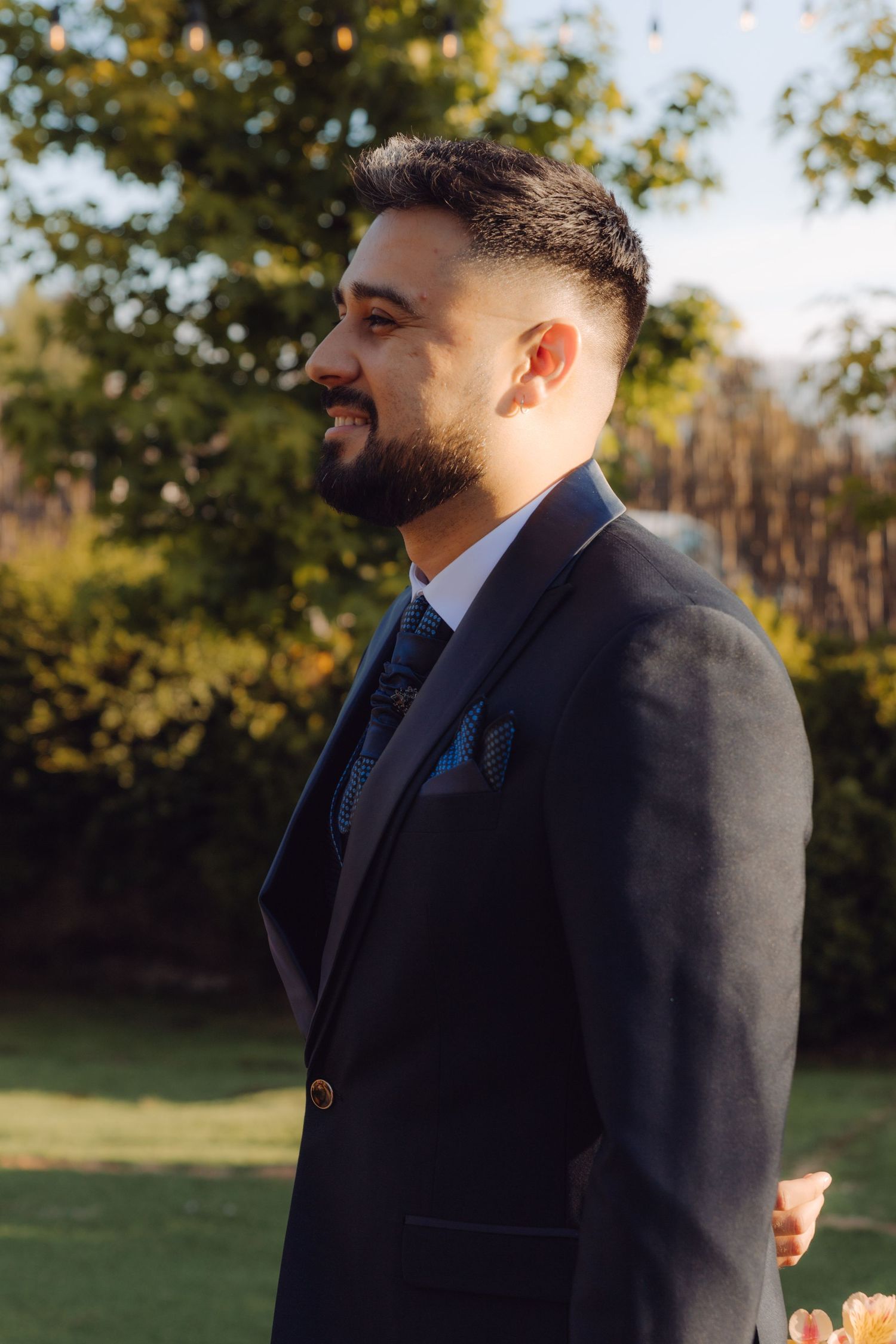 Handsome groom in a dark suit smiles outdoors during golden hour at a wedding ceremony.