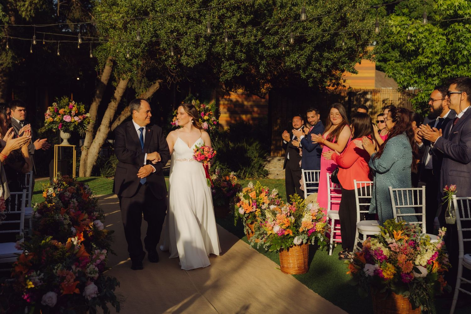 Smiling bride and her father walk joyfully down the aisle as wedding guests celebrate at an outdoor ceremony.