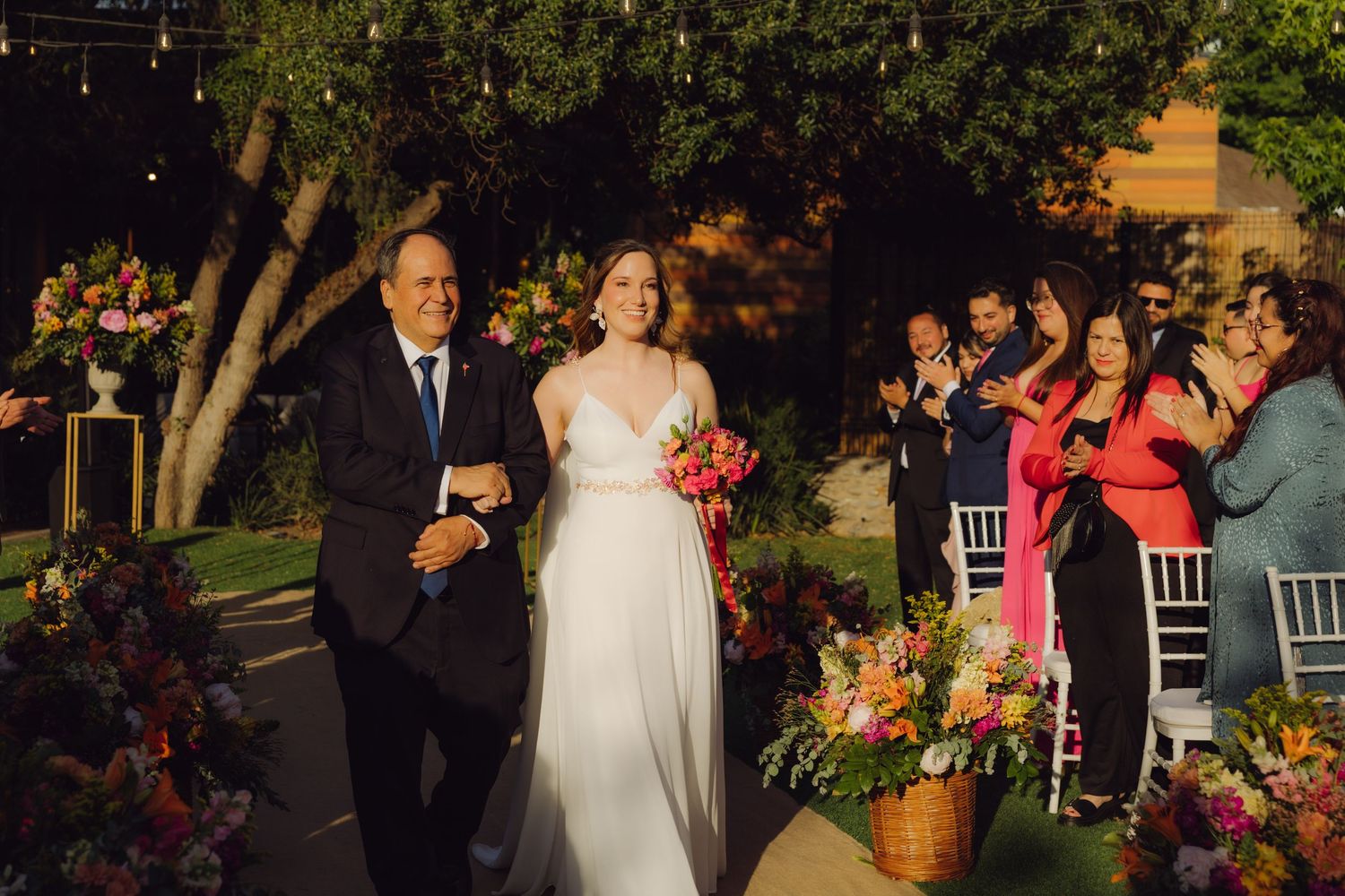 Bride walks down the aisle with her father as guests applaud at an outdoor evening wedding ceremony.