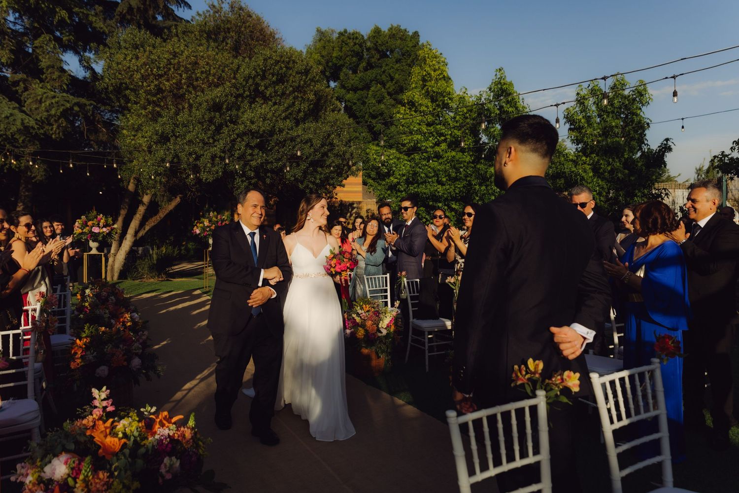 Bride walks down the aisle with escort at outdoor wedding ceremony surrounded by colorful floral arrangements.