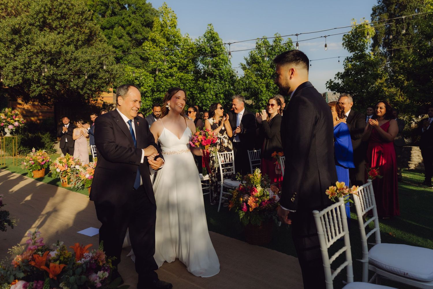 Father escorts bride down the aisle toward groom at elegant outdoor garden wedding ceremony.
