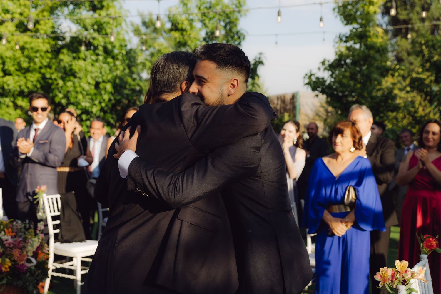 Groom and father of the bride share an emotional hug during outdoor wedding ceremony.