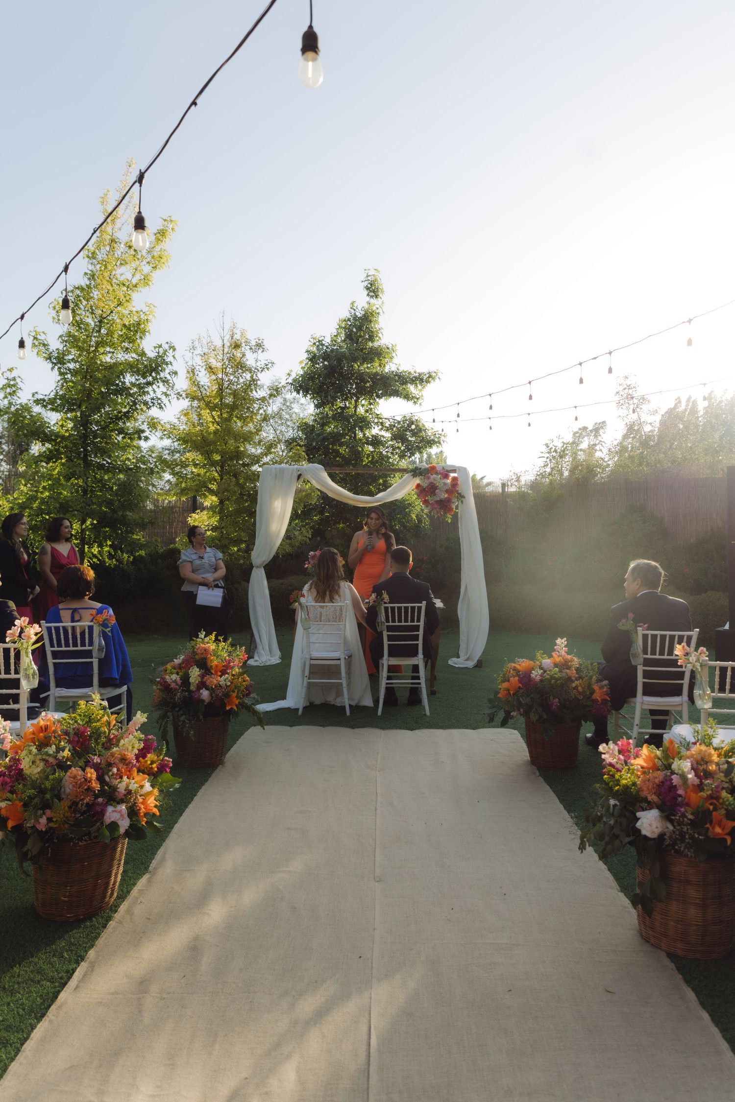 Elegant outdoor wedding ceremony aisle with floral arrangements, string lights, and draped arch at sunset.