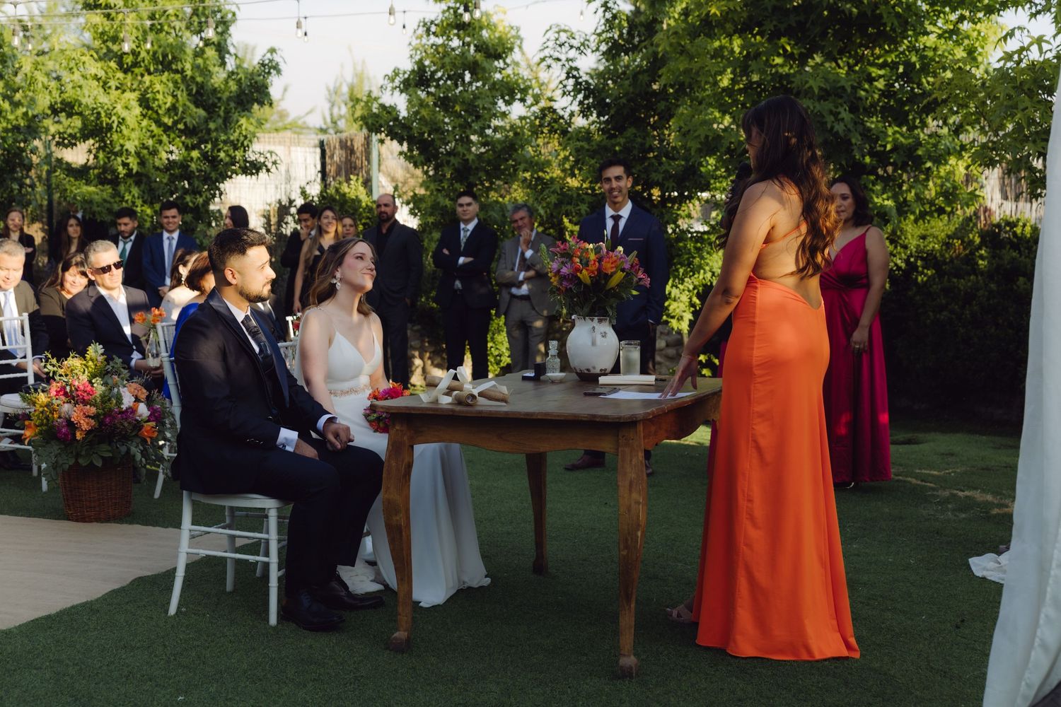Bride and groom seated at wooden table during outdoor wedding ceremony with officiant in orange dress.