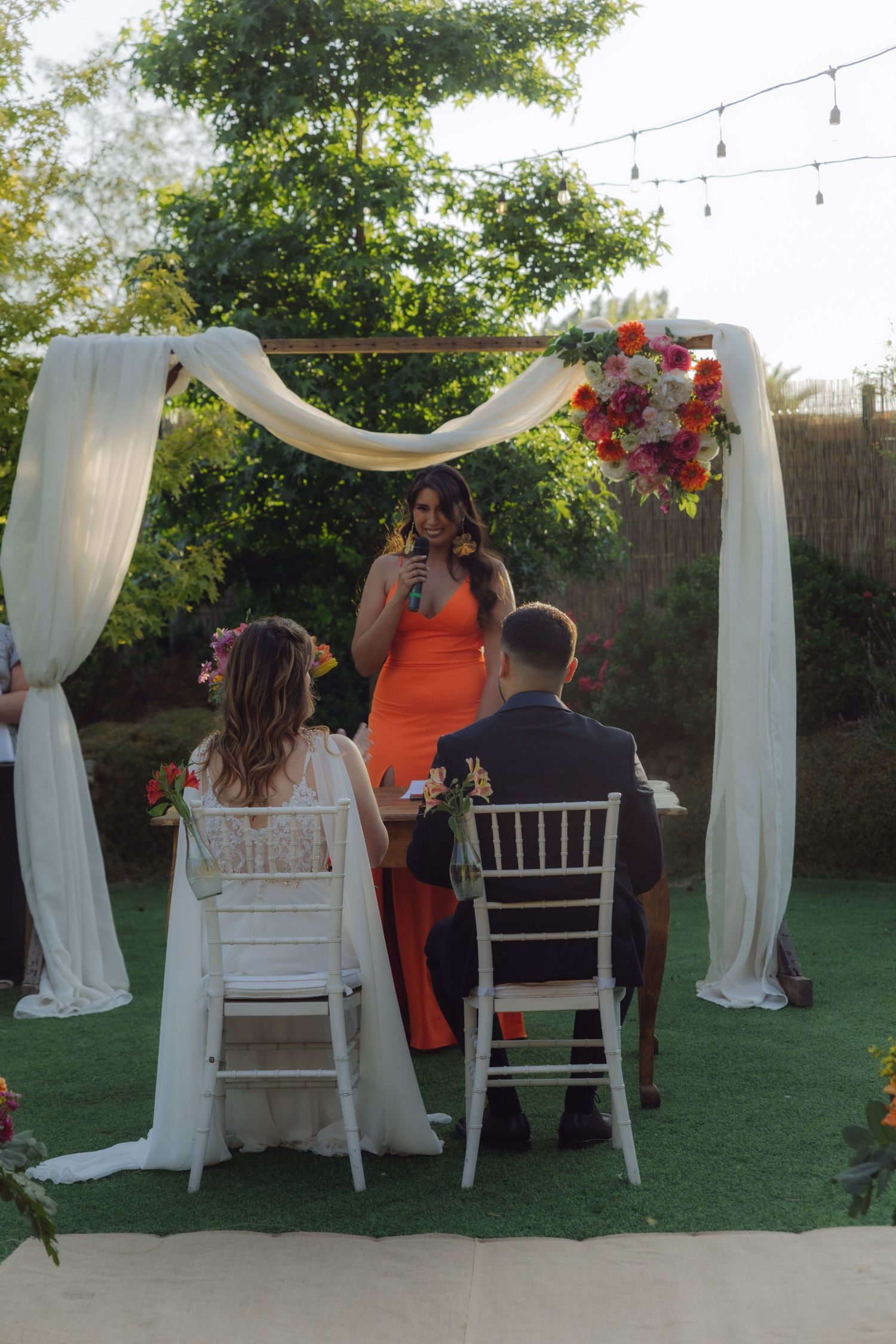 Officiant in orange dress speaks at outdoor wedding ceremony under draped floral arch with string lights.