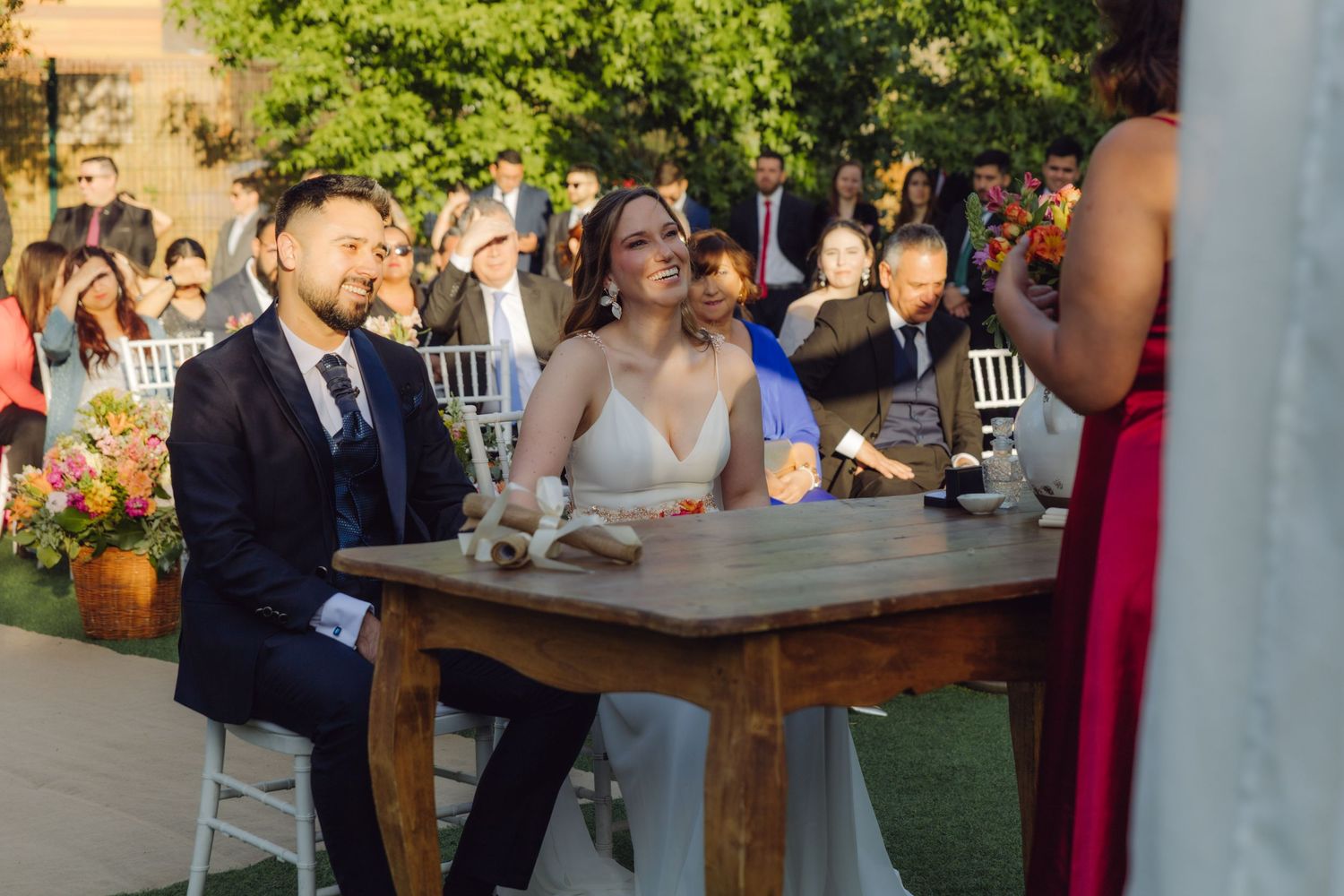 Bride and groom smile joyfully while seated at wooden table during outdoor wedding ceremony.