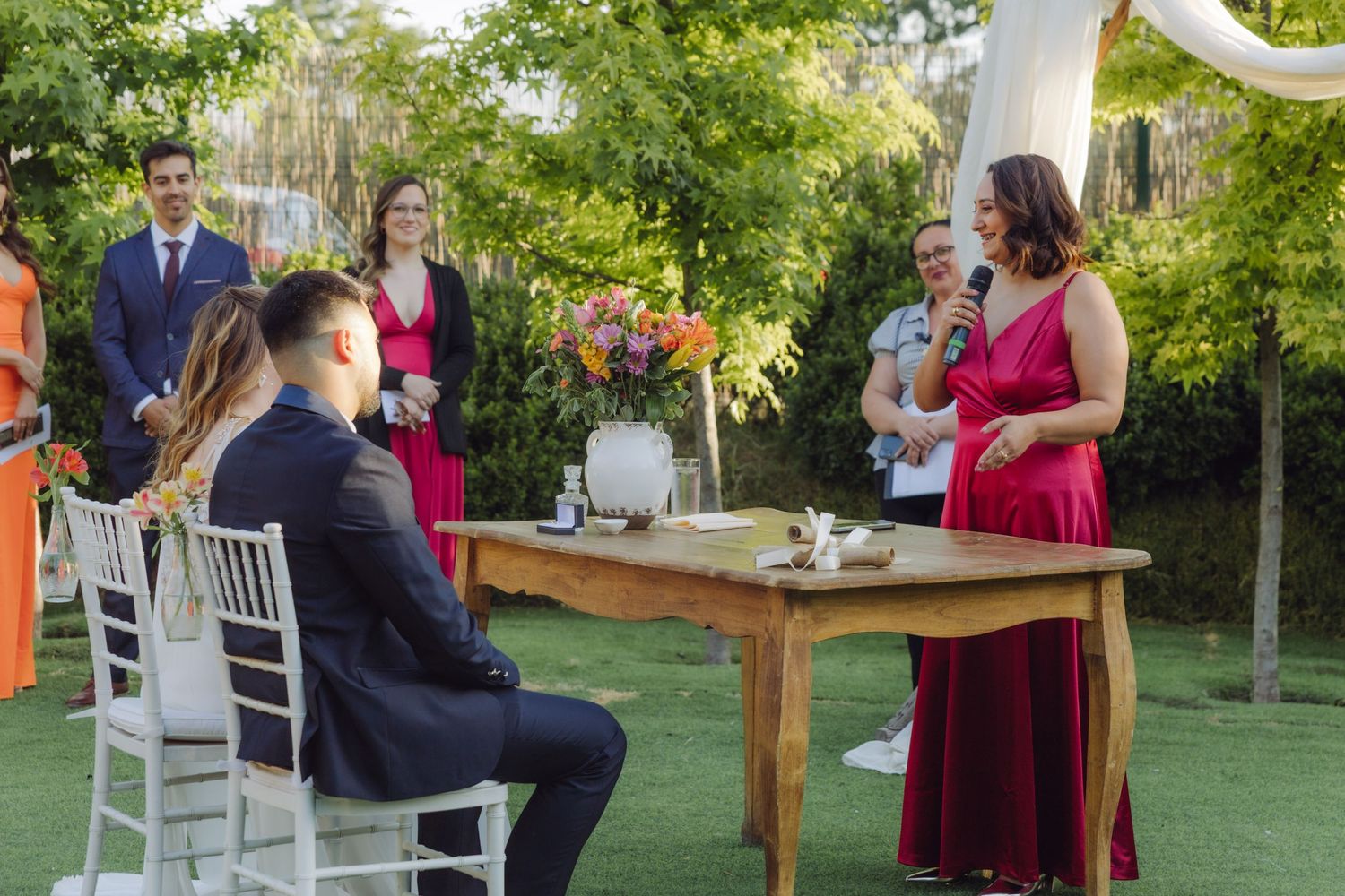 Officiant in red dress addresses bride and groom seated at rustic wooden table at outdoor ceremony.
