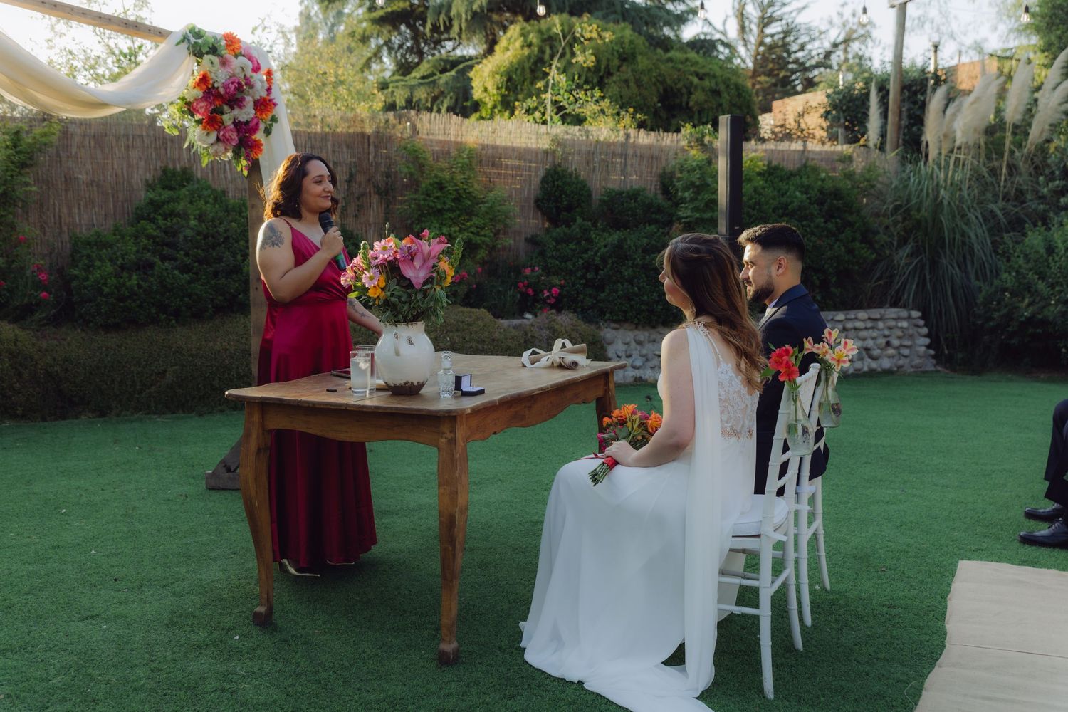 Officiant in burgundy dress speaks at wooden altar during outdoor garden wedding ceremony with seated bride and groom.
