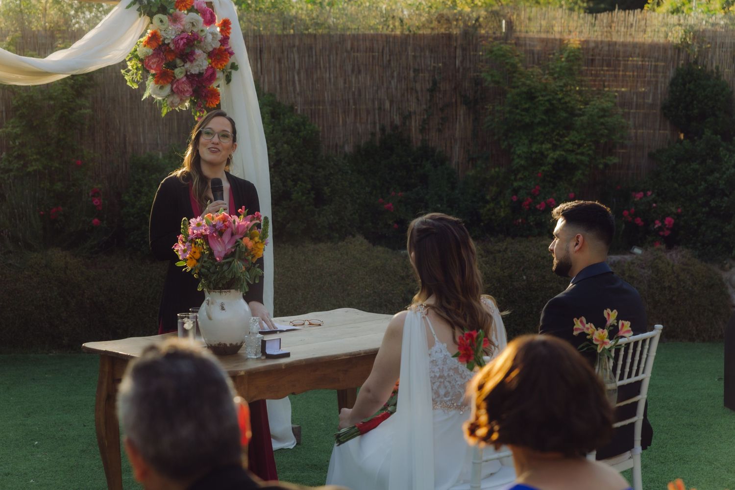 Wedding officiant addresses seated couple at flower-adorned wooden table during intimate outdoor garden ceremony at dusk.