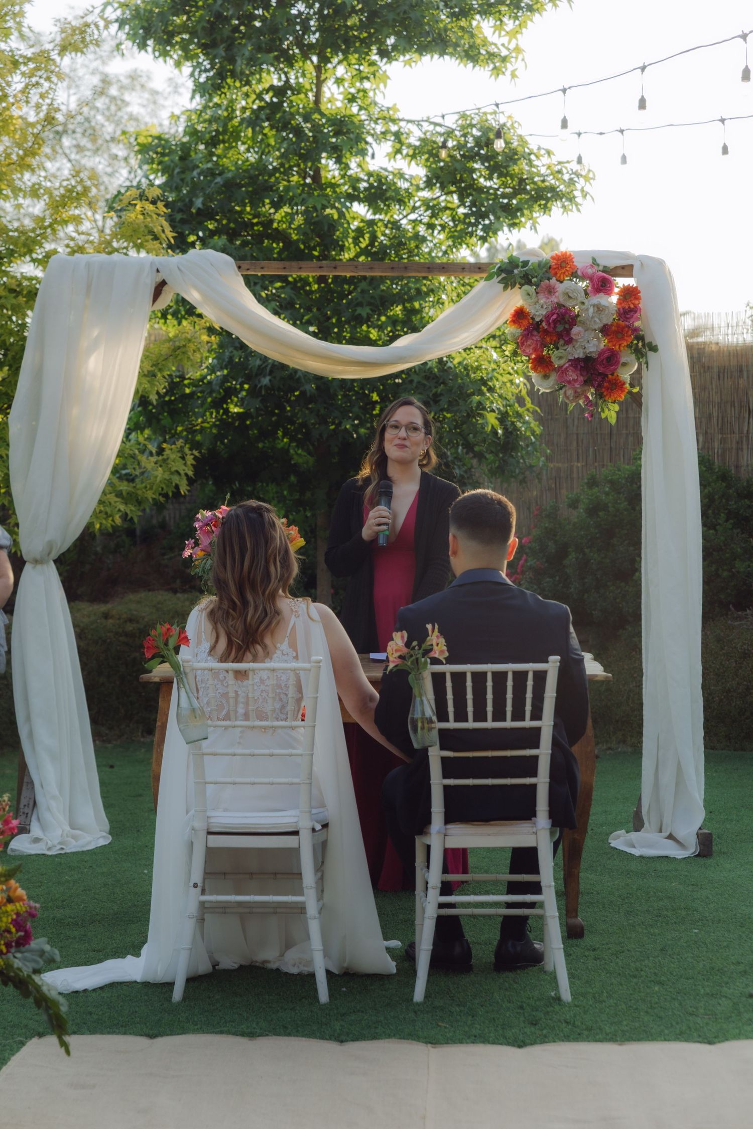 Bride and groom seated before decorated arch with white draping and colorful flowers during outdoor wedding ceremony.