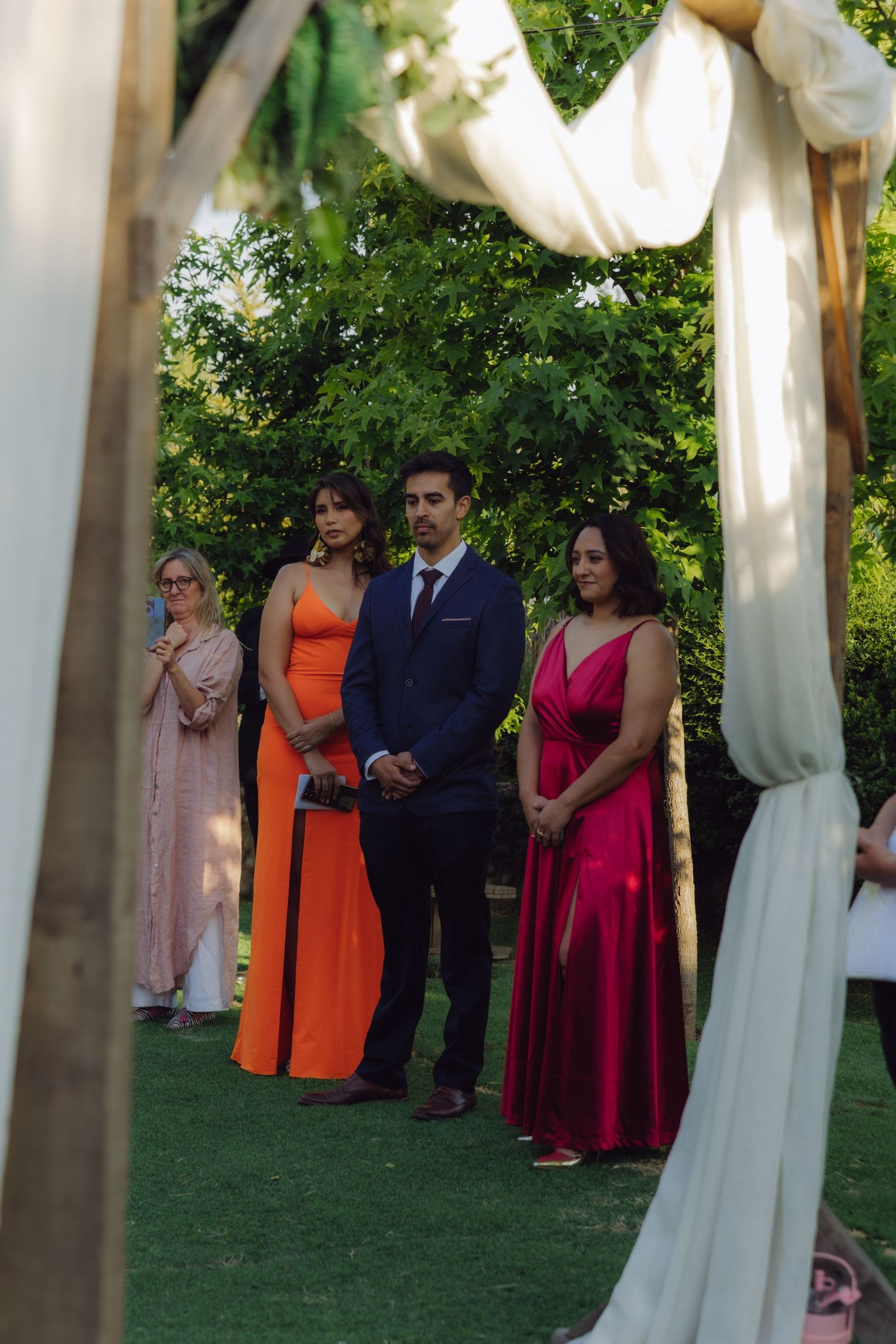 Wedding guests in elegant orange and red gowns stand attentively watching outdoor garden ceremony through decorated arch.