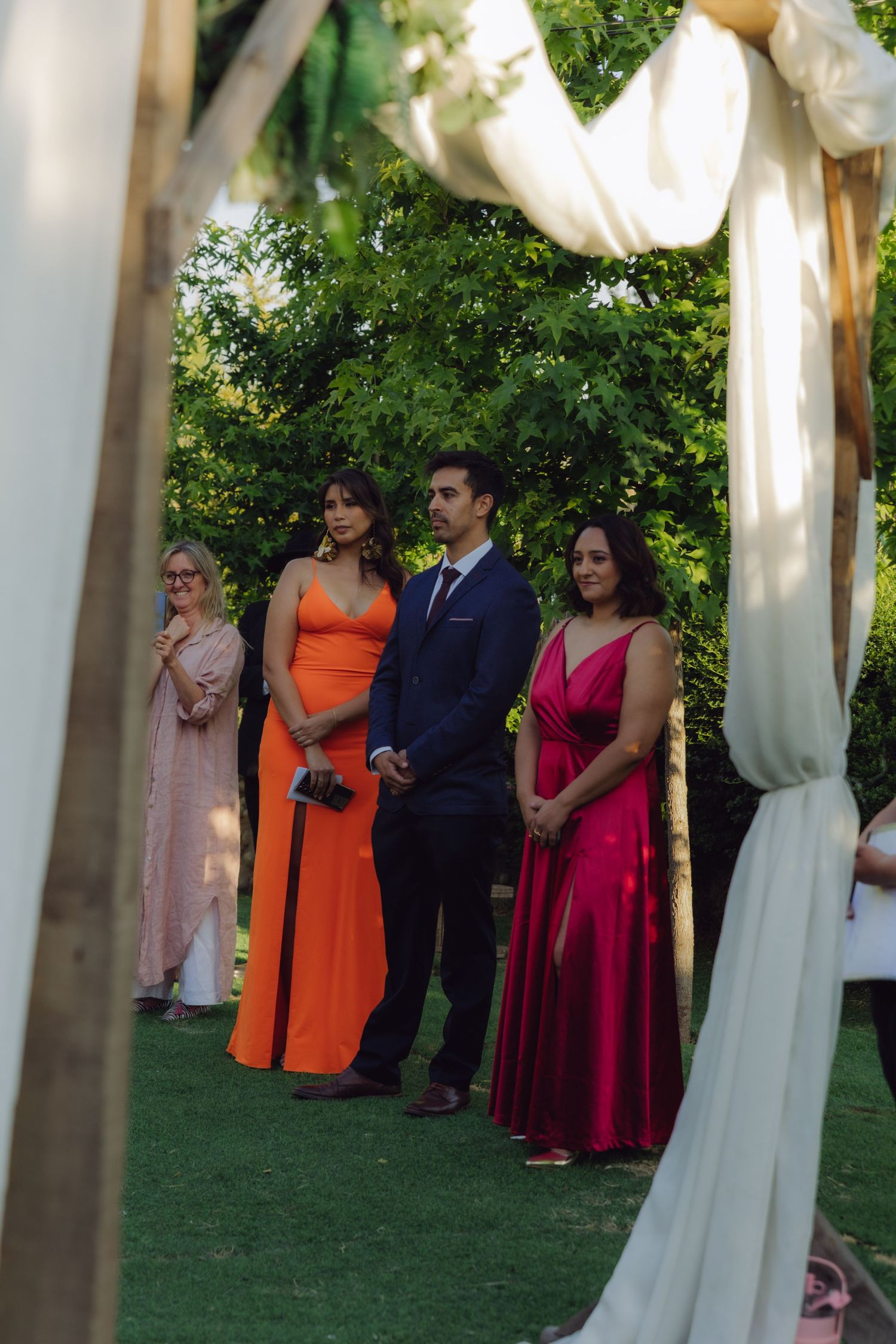 Formally dressed wedding guests in vibrant orange and red gowns observe outdoor garden ceremony through floral arch frame.