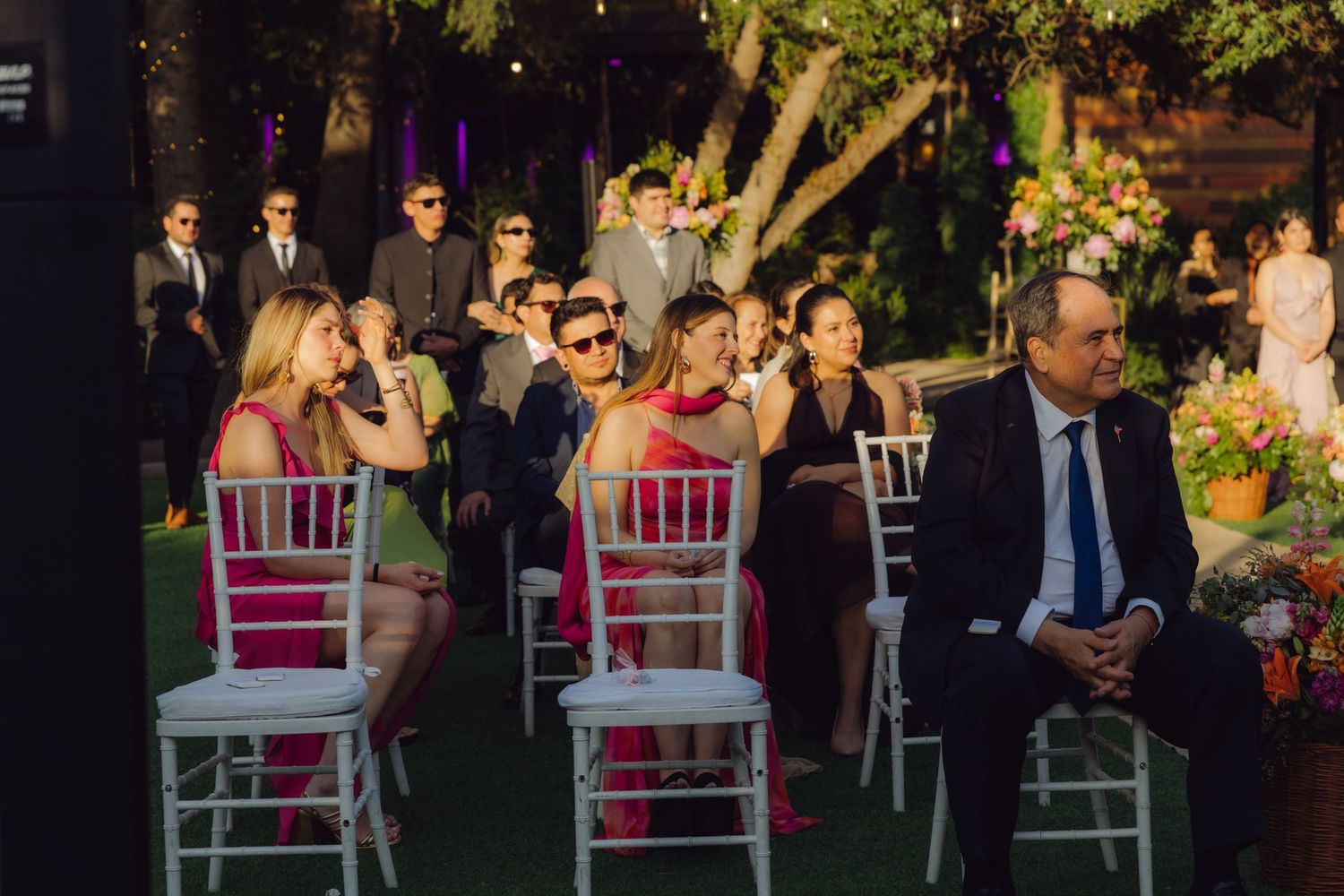 Wedding guests seated in white chiavari chairs watch evening outdoor ceremony in garden with string lights overhead.