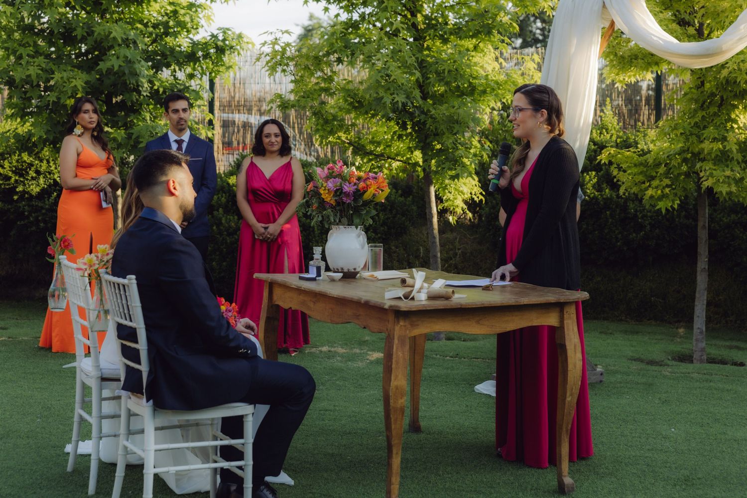 Wedding officiant in burgundy speaks to seated groom at outdoor ceremony with bridal party standing in background.