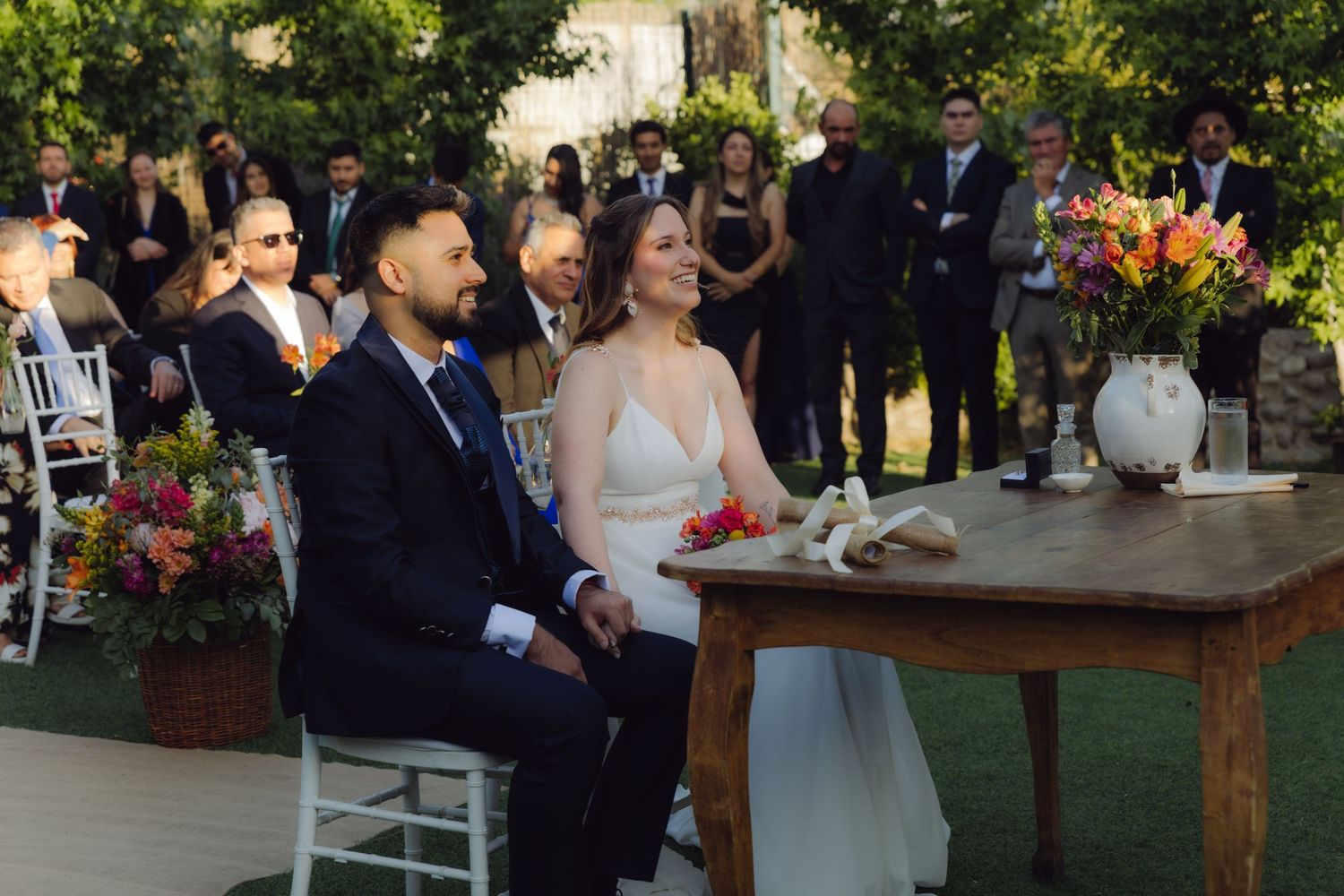 Smiling bride and groom sit together at wooden table during outdoor garden wedding ceremony with guests behind them.