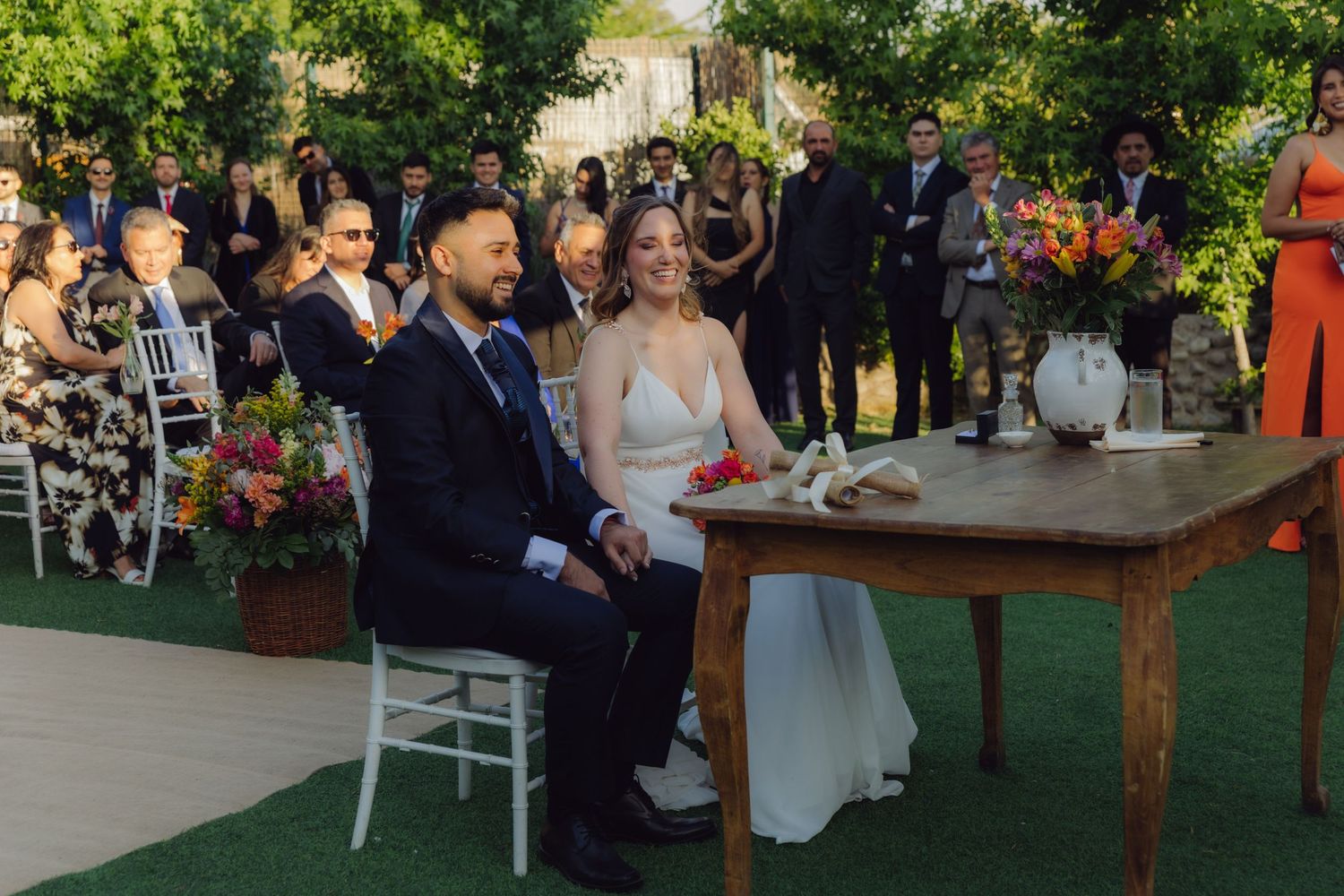Happy bride and groom seated at ceremonial table smile during outdoor wedding with guests gathered in background.