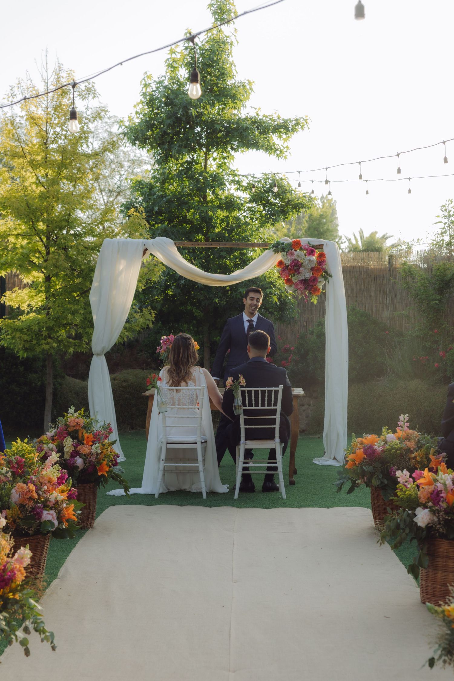 Wide view of outdoor garden wedding ceremony with couple seated under white draped arch lined with string lights.