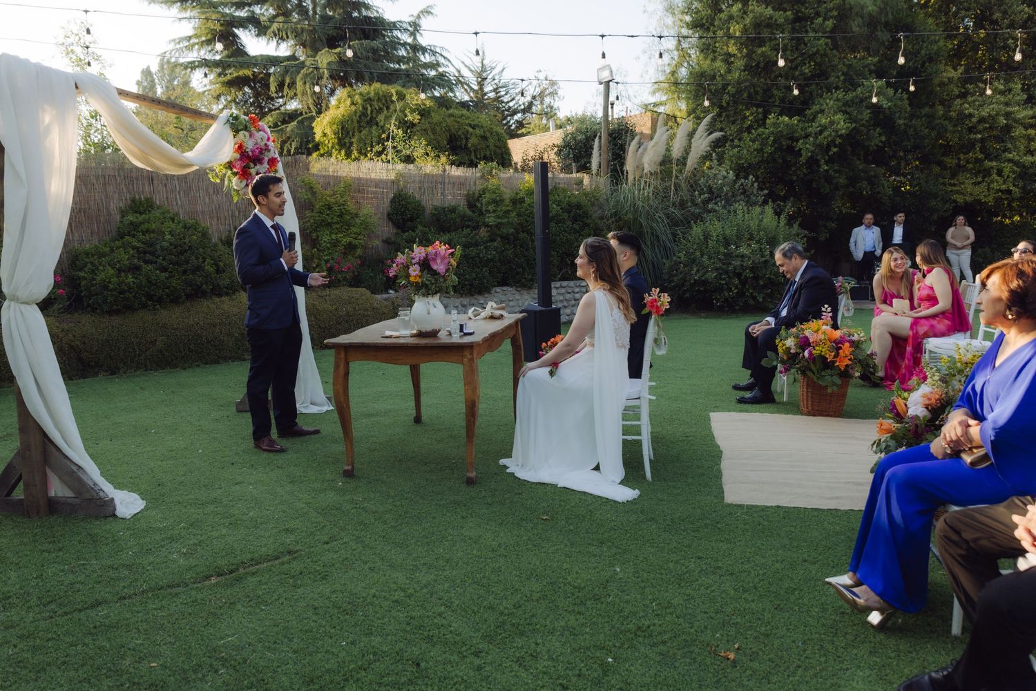 Outdoor wedding ceremony on green lawn with bride and groom at wooden altar under draped arch with colorful floral arrangements.