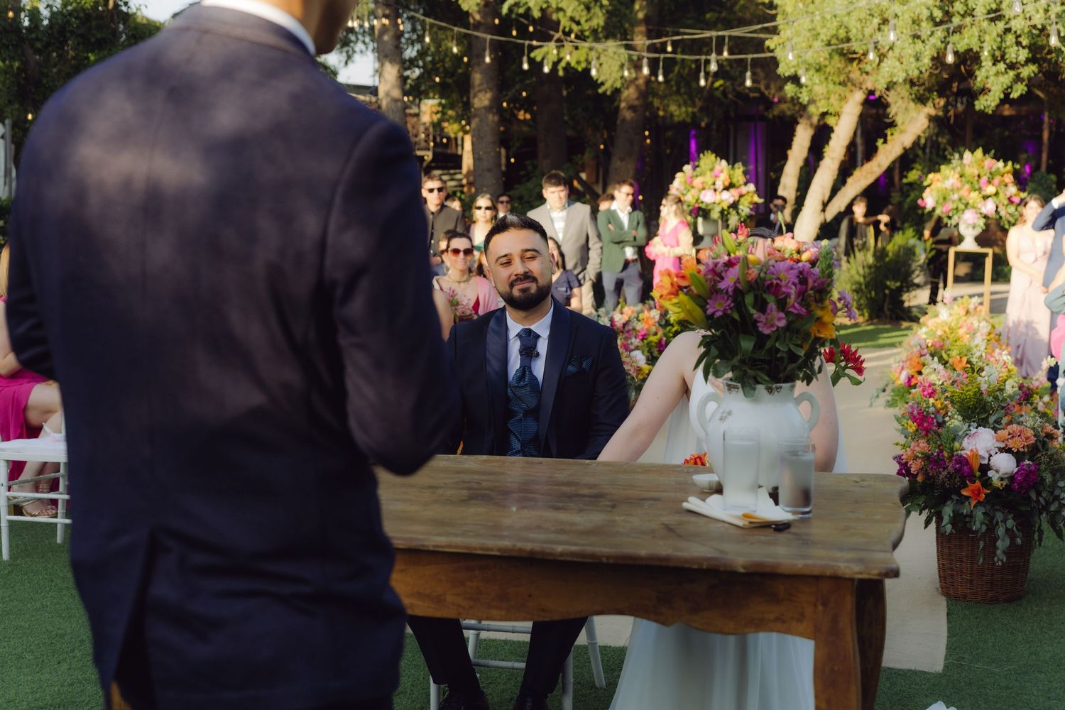 Groom gazes lovingly at bride during outdoor wedding ceremony at rustic wooden table adorned with vibrant floral bouquets.