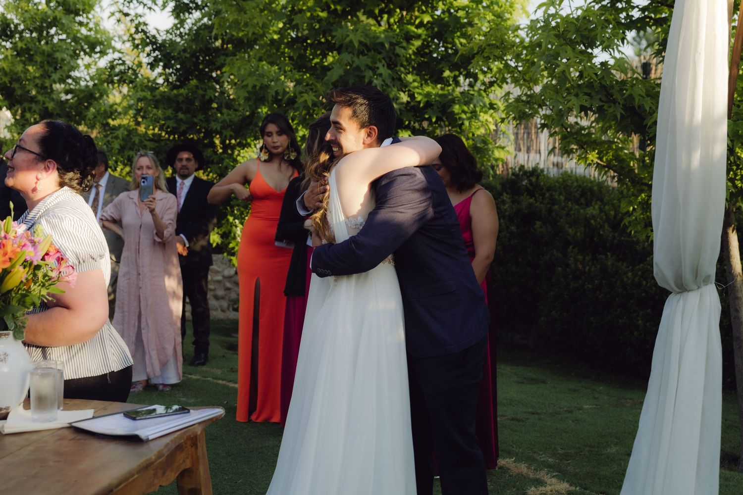 Bride and groom share a heartfelt hug at outdoor wedding ceremony surrounded by guests in colorful formal attire.