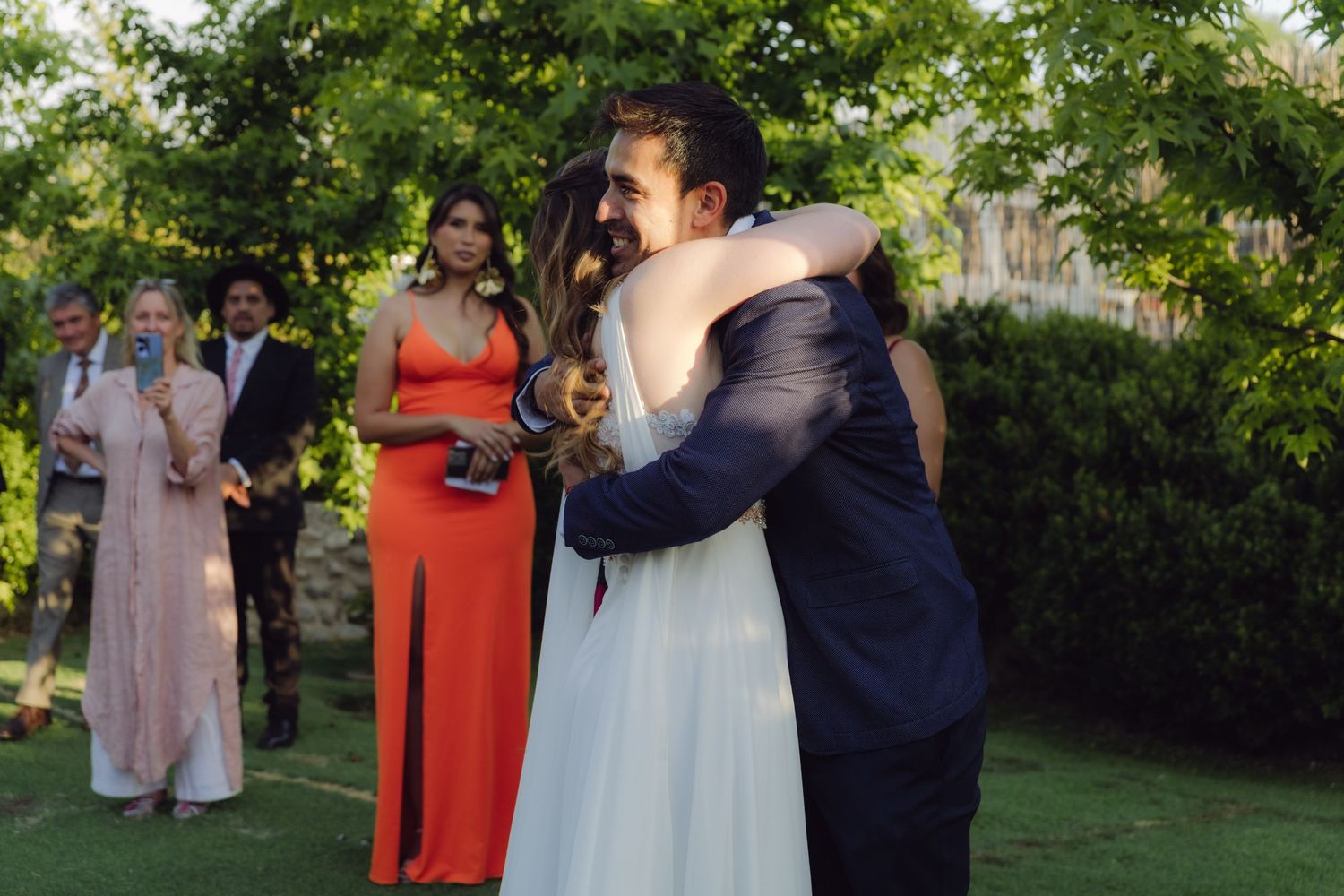 Newlyweds embrace joyfully after outdoor garden wedding ceremony while guests in elegant attire look on and celebrate.