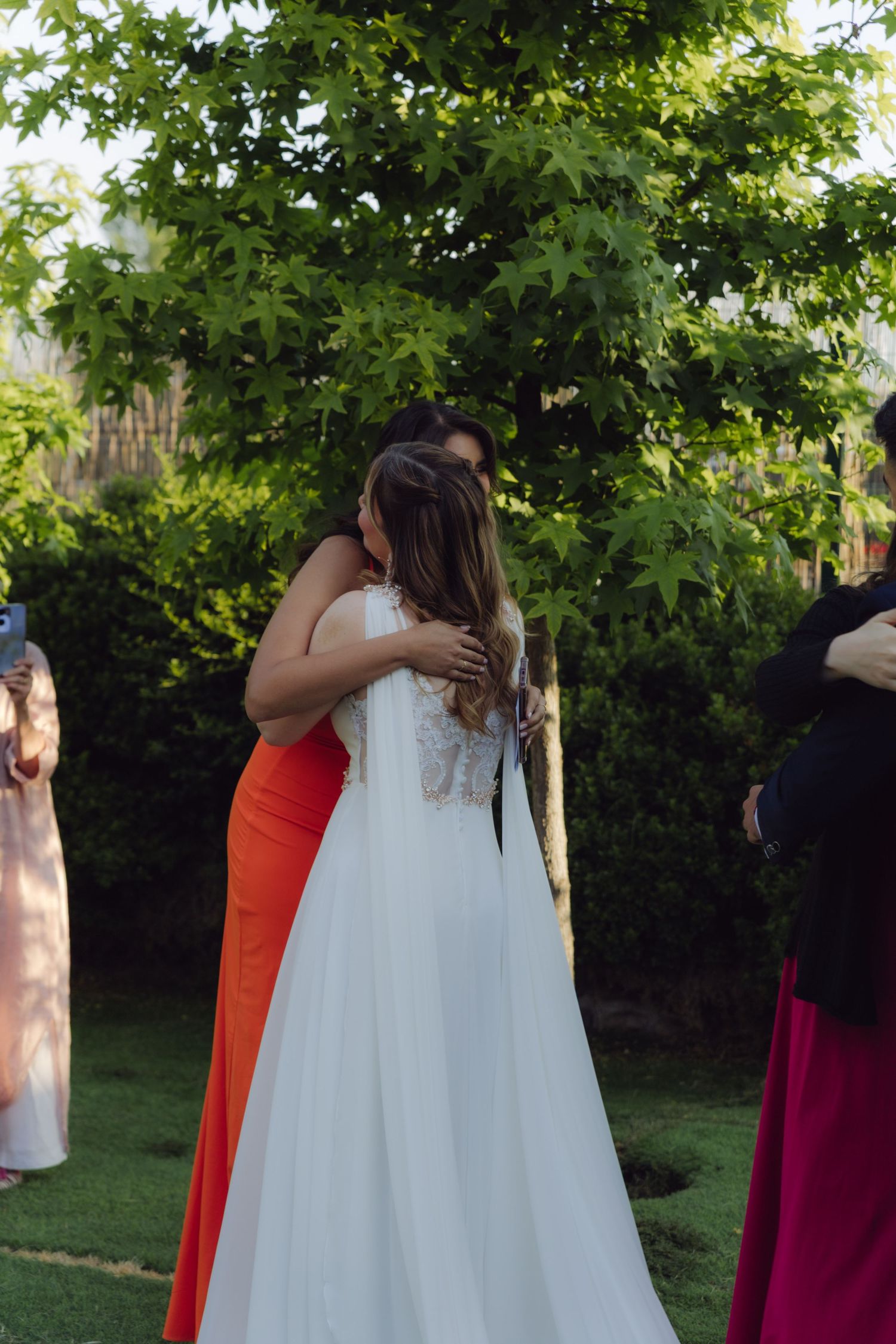 Bride in white lace gown shares warm embrace with guest in orange dress during outdoor garden wedding celebration.