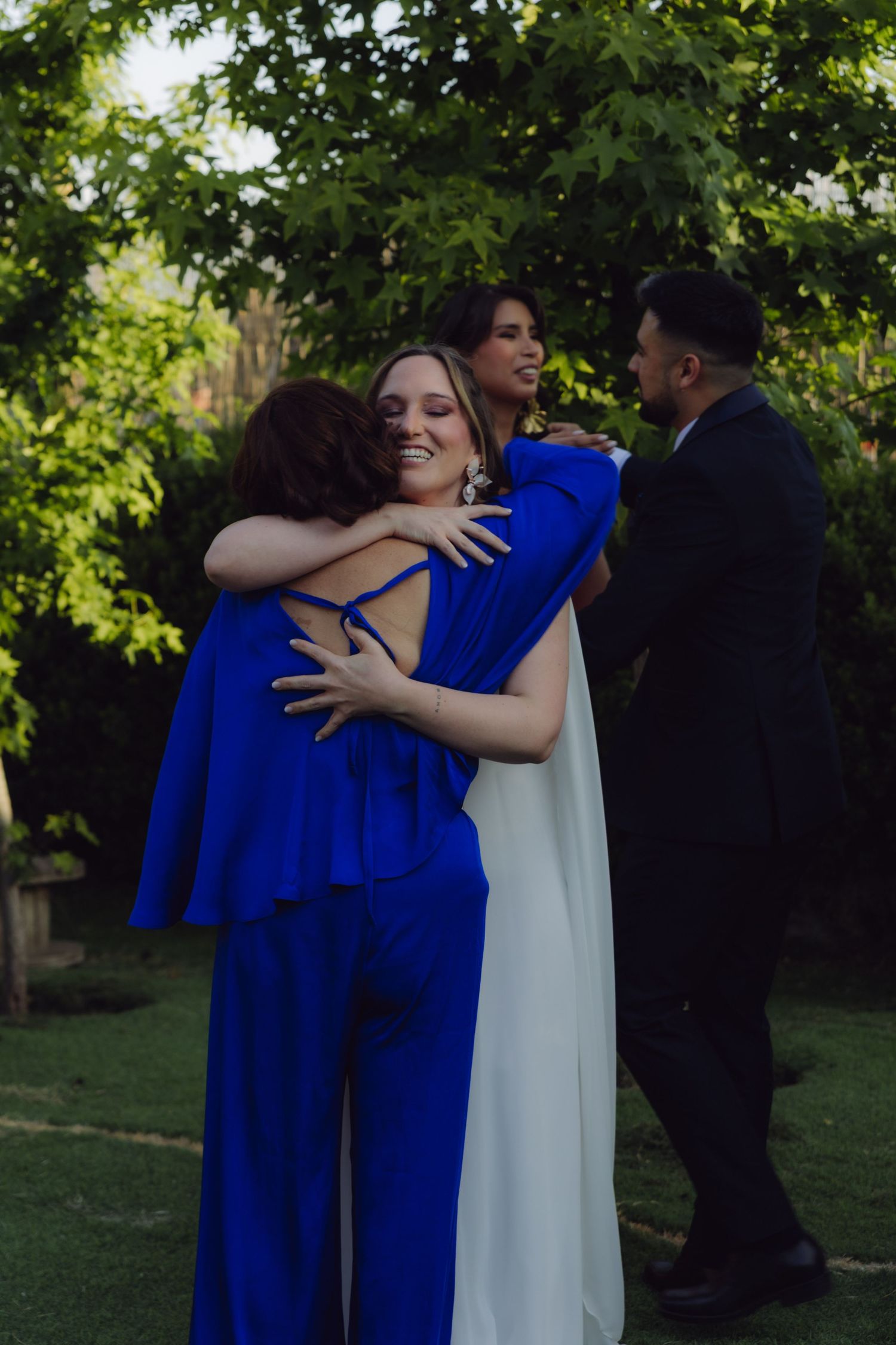 Wedding guests in royal blue and white outfits share joyful hugs in lush green garden after ceremony.