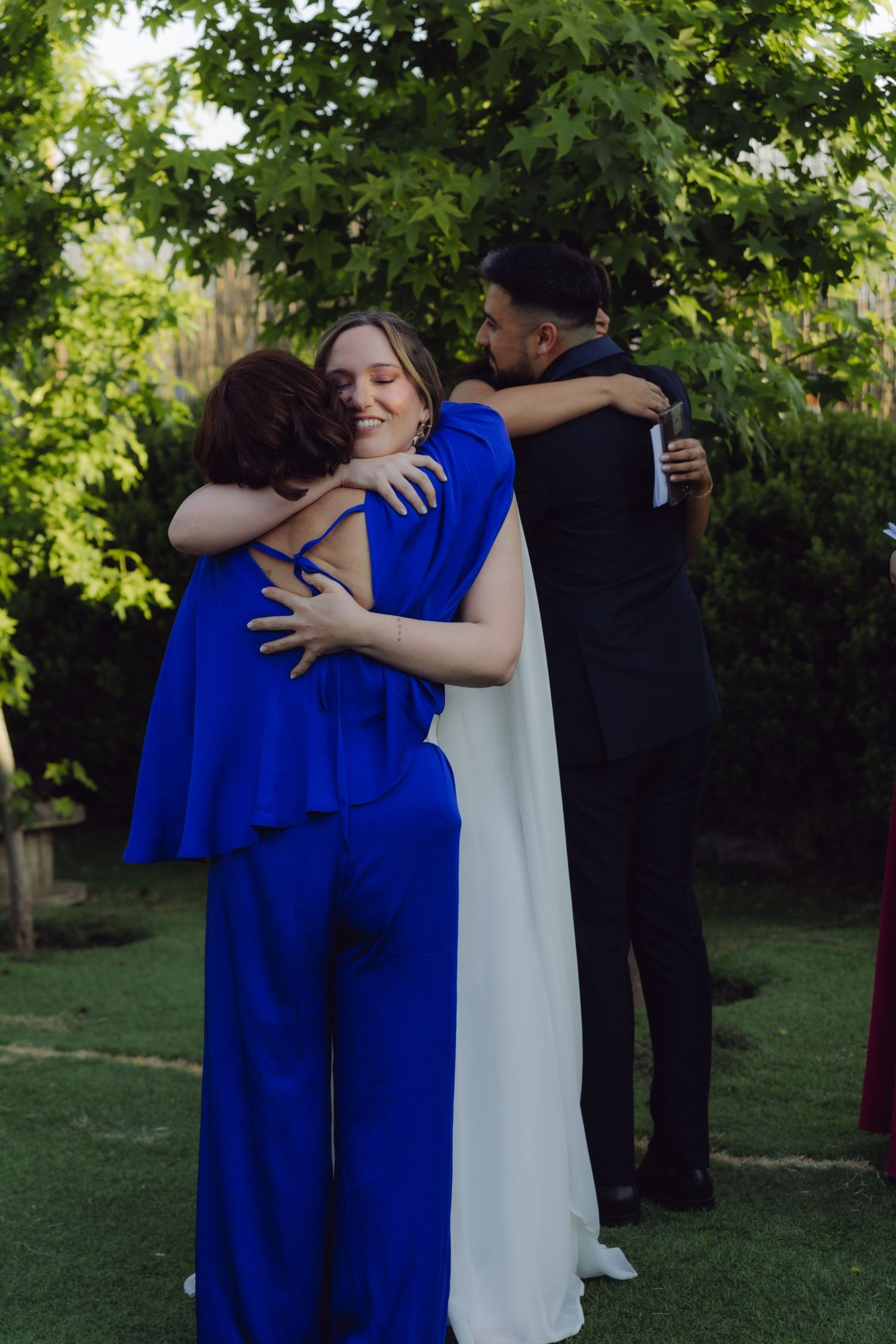 Bride in white gown and guest in royal blue embrace warmly while groom hugs another guest in garden.