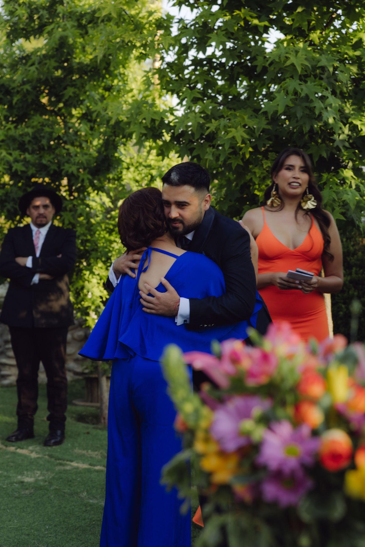 Groom in dark suit embraces guest in royal blue outfit at outdoor wedding reception surrounded by greenery.