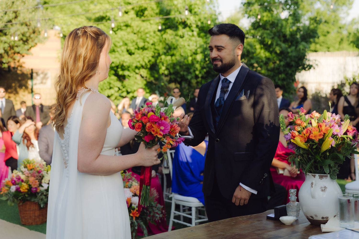 Bride holding vibrant bouquet faces smiling groom at outdoor wedding ceremony with colorful floral arrangements on table.
