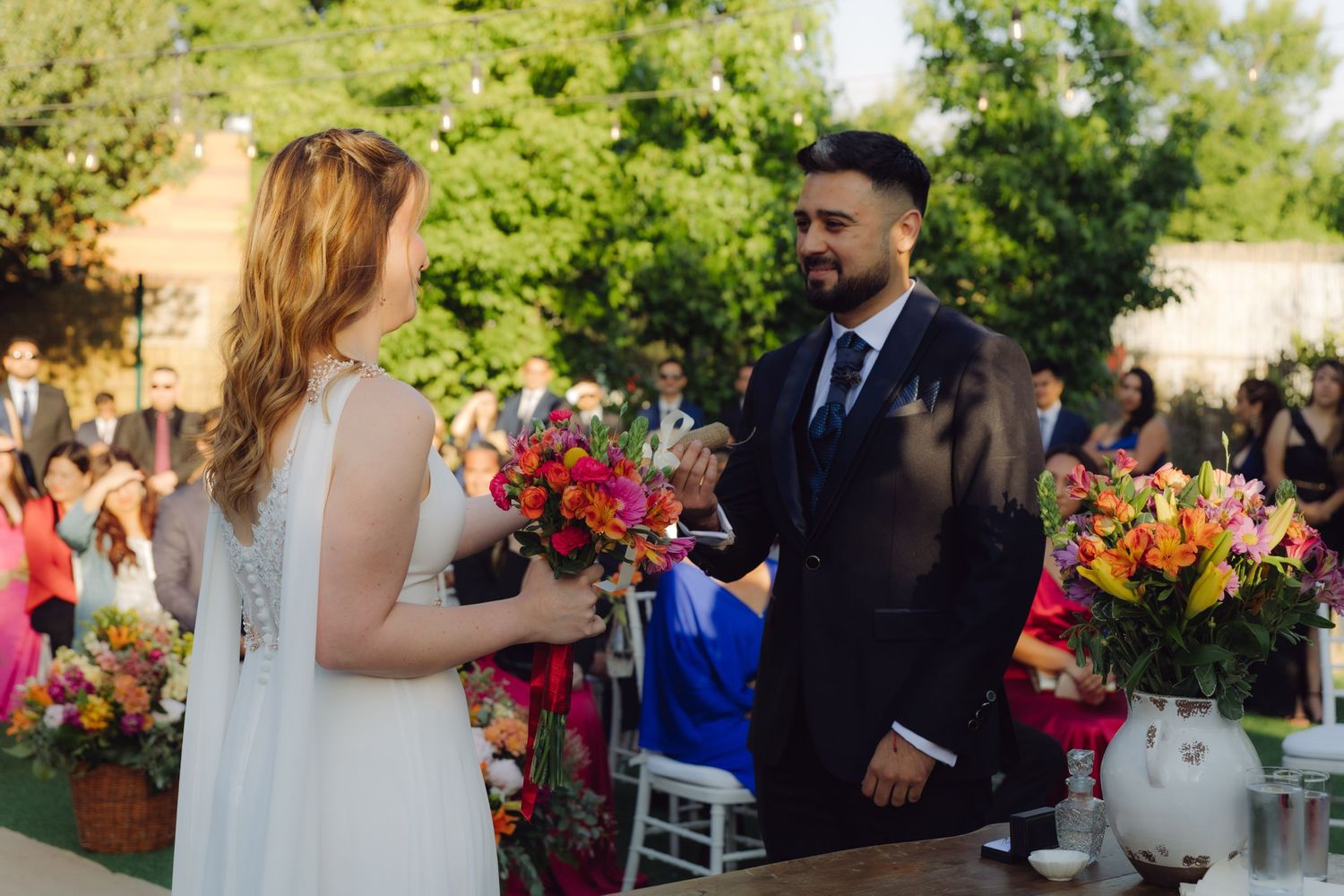 Bride and groom exchange vows at sunlit outdoor wedding ceremony with bright tropical floral arrangements on wooden table.